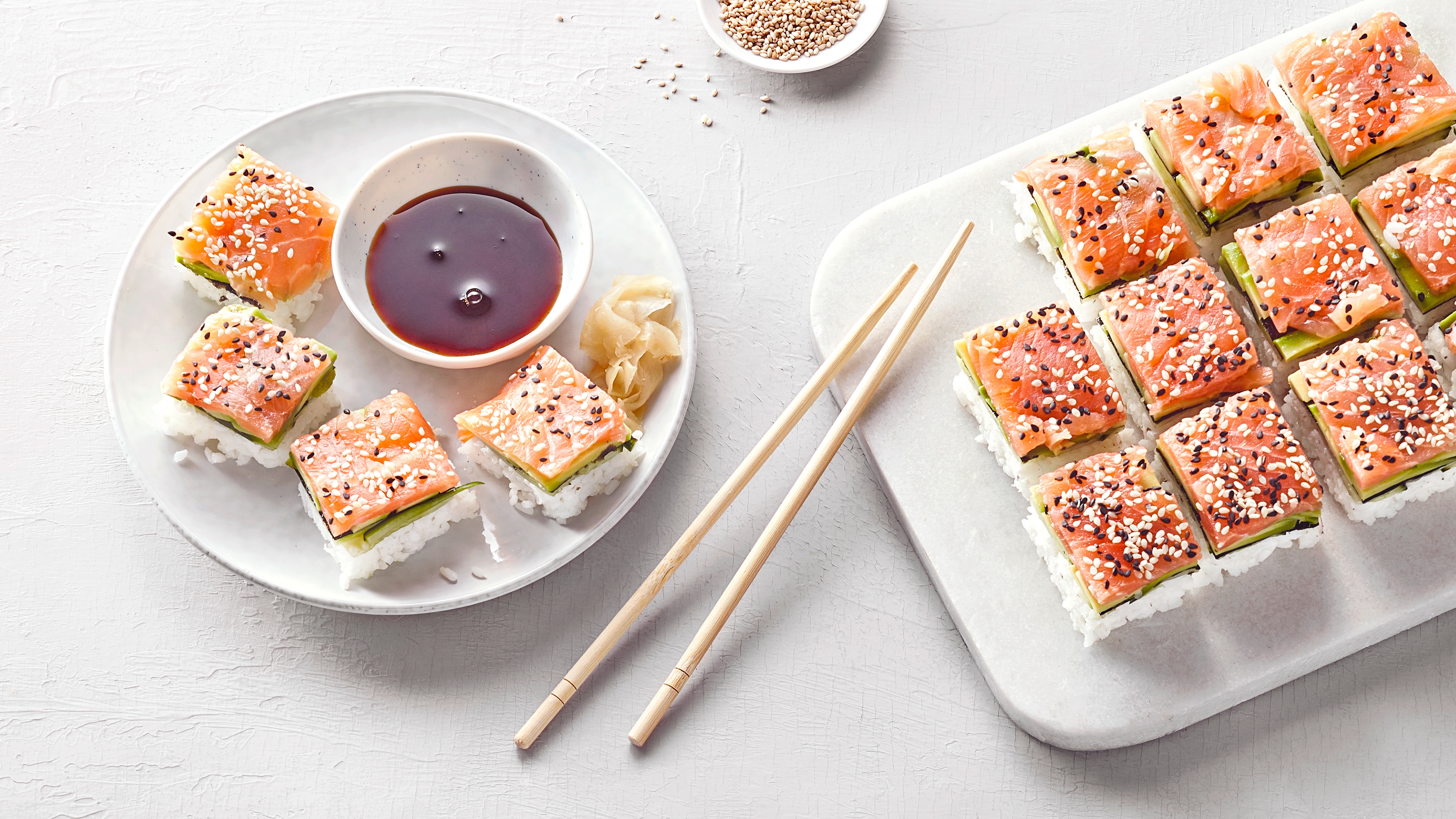 Blech mit mehreren quadratischen Sushi-Stücken aus Lachs, Avocado, Reis und Sesam. Links daneben ein Teller mit vier Sushi-Stücken und einer süß-sauren Soße.