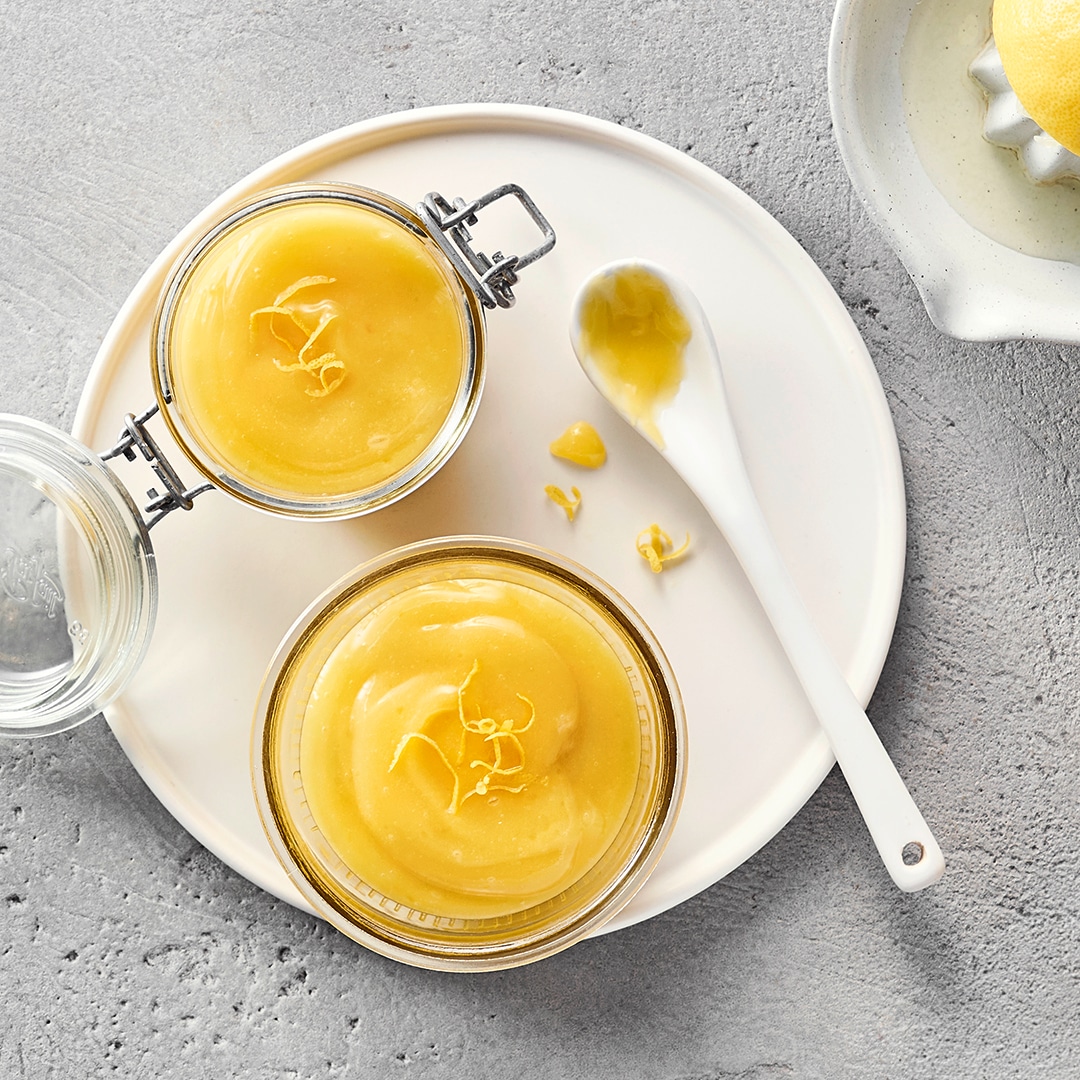 Zwei Gläsern gelber Creme, garniert mit feinen Zitronenzesten und einem weißen Löffel mit etwas Creme darauf.