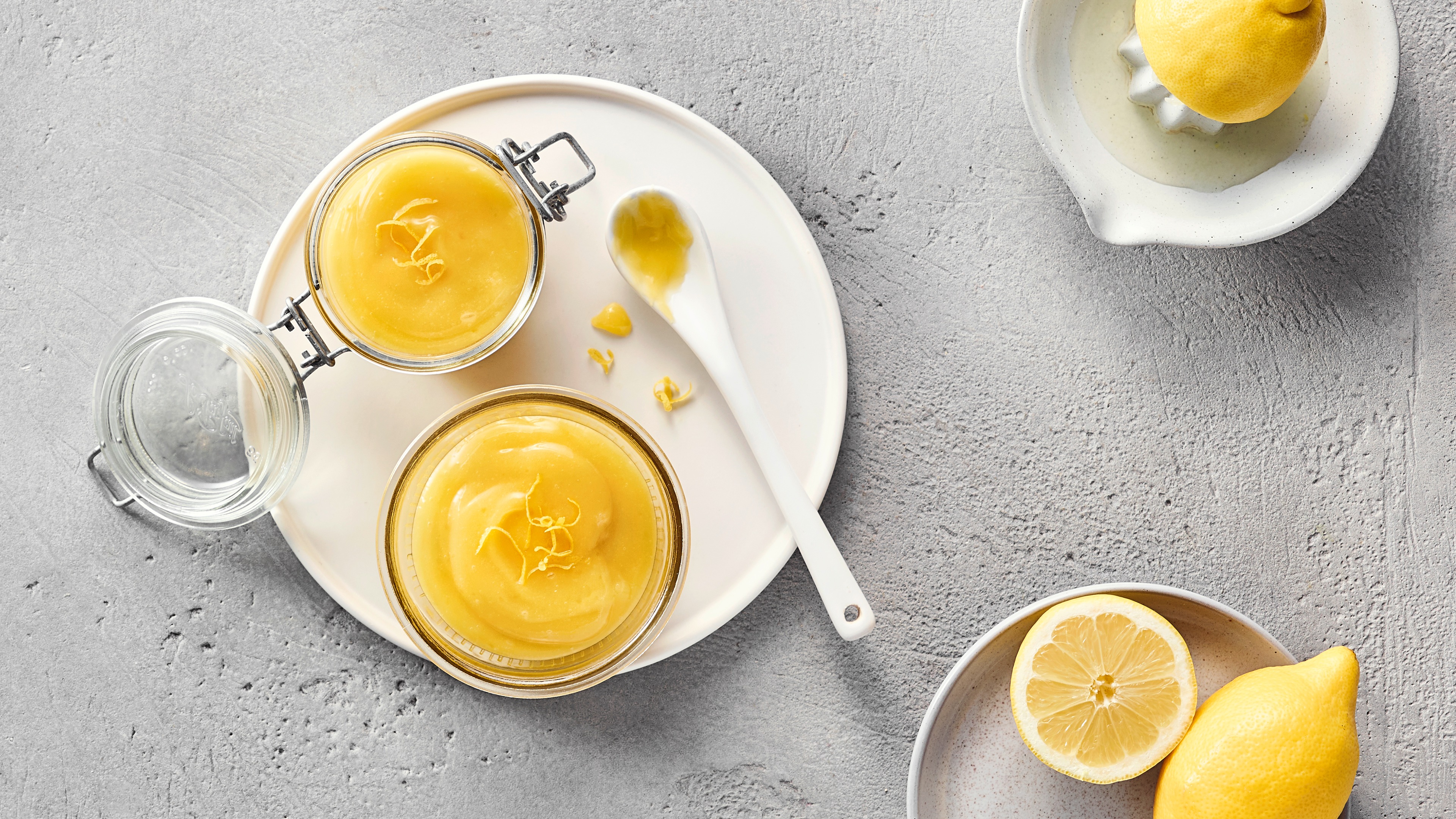 Zwei Gläsern gelber Creme, garniert mit feinen Zitronenzesten und einem weißen Löffel mit etwas Creme darauf.
