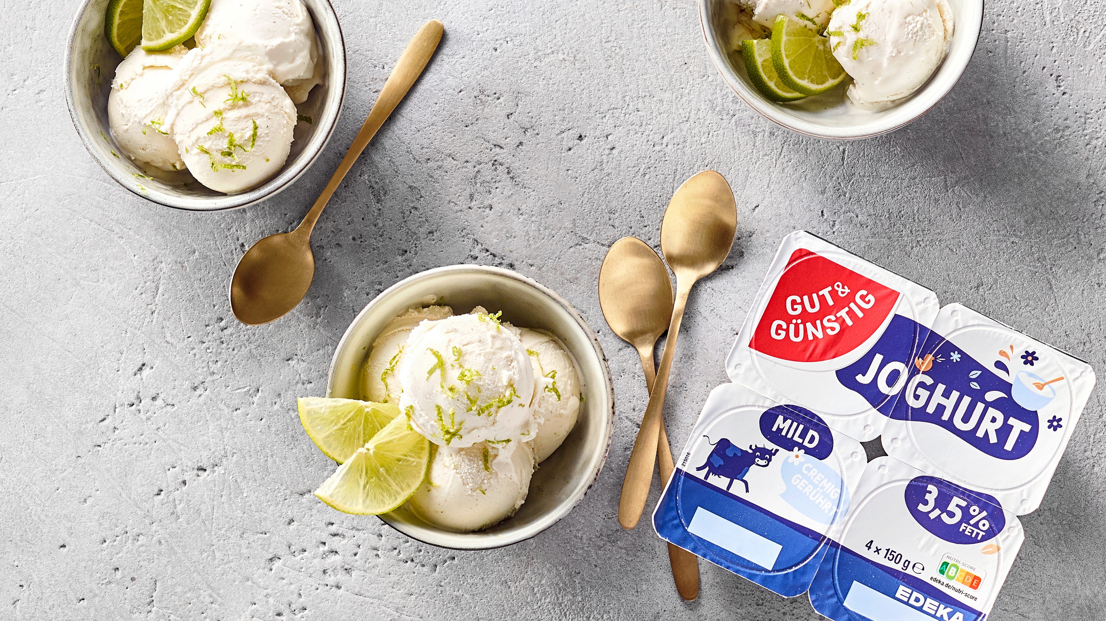 Schalen mit Limetten-Joghurteis, garniert mit Limettenspalten; daneben GUT&GÜNSTIG Joghurtbecher und goldene Löffel.