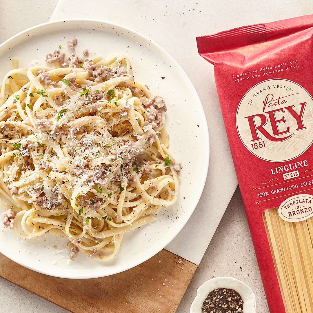 Weißer Teller mit dünnen langen Nudeln, heller Soße und geriebenem Käse. Daneben eine Packung Pasta Rey Linguine