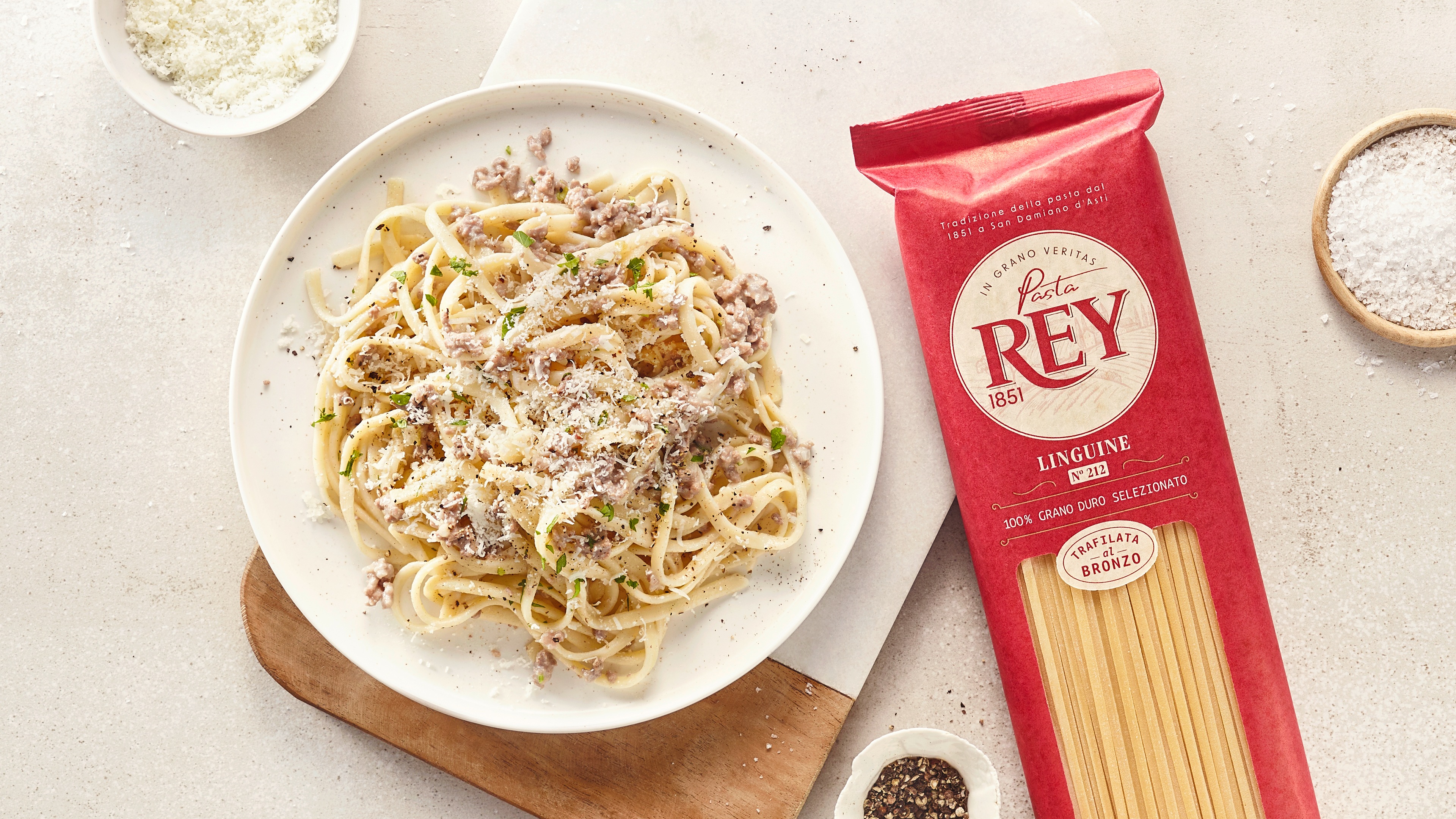 Weißer Teller mit dünnen langen Nudeln, heller Soße und geriebenem Käse. Daneben eine Packung Pasta Rey Linguine