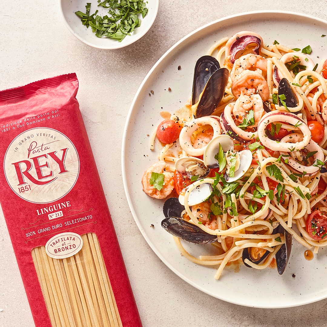 Linguine mit Meeresfrüchten, Muscheln, Garnelen, Tomaten und frischen Kräutern auf einem weißen Teller; daneben Pasta Rey Linguine Verpackung.