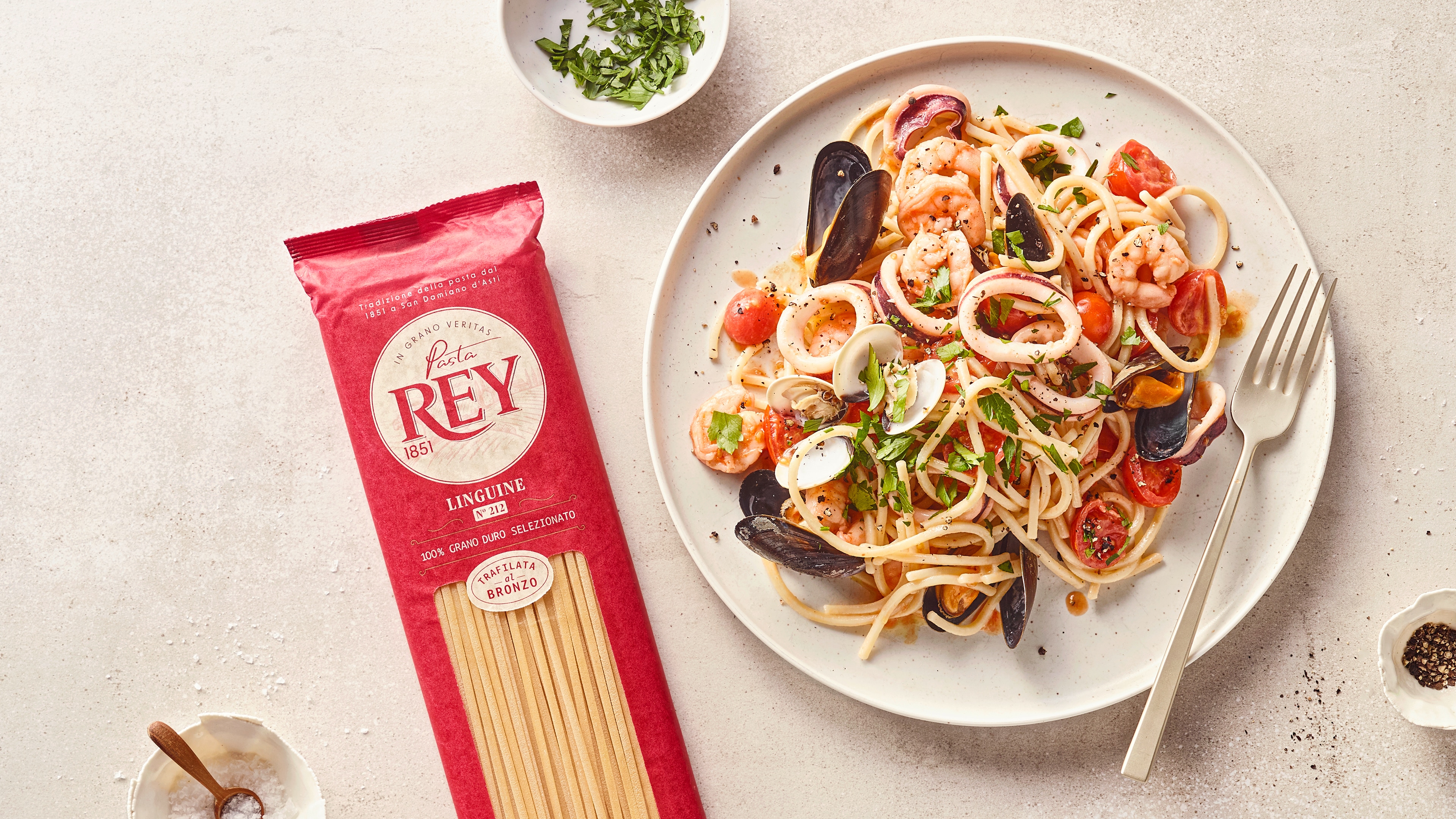 Linguine mit Meeresfrüchten, Muscheln, Garnelen, Tomaten und frischen Kräutern auf einem weißen Teller; daneben Pasta Rey Linguine Verpackung.