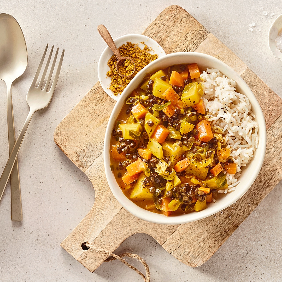 Eine Schüssel mit weißem Reis, gelben Würfeln, orangefarbenen Würfeln und braunen Linsen in gelber Soße.