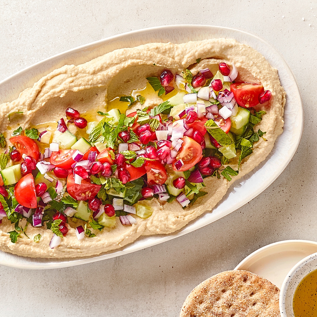 Große Platte mit Hummus und darauf Tomatenhälften, Gurkenscheiben, Granatapfelkernen und Zwiebelstückchen