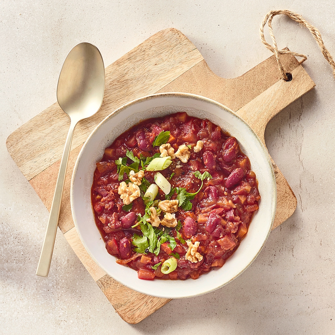 Eine Schüssel mit roter Soße, darin rote Bohnen und kleine Stücke, darauf grüne Kräuter, helle Walnüsse und Lauchstücke