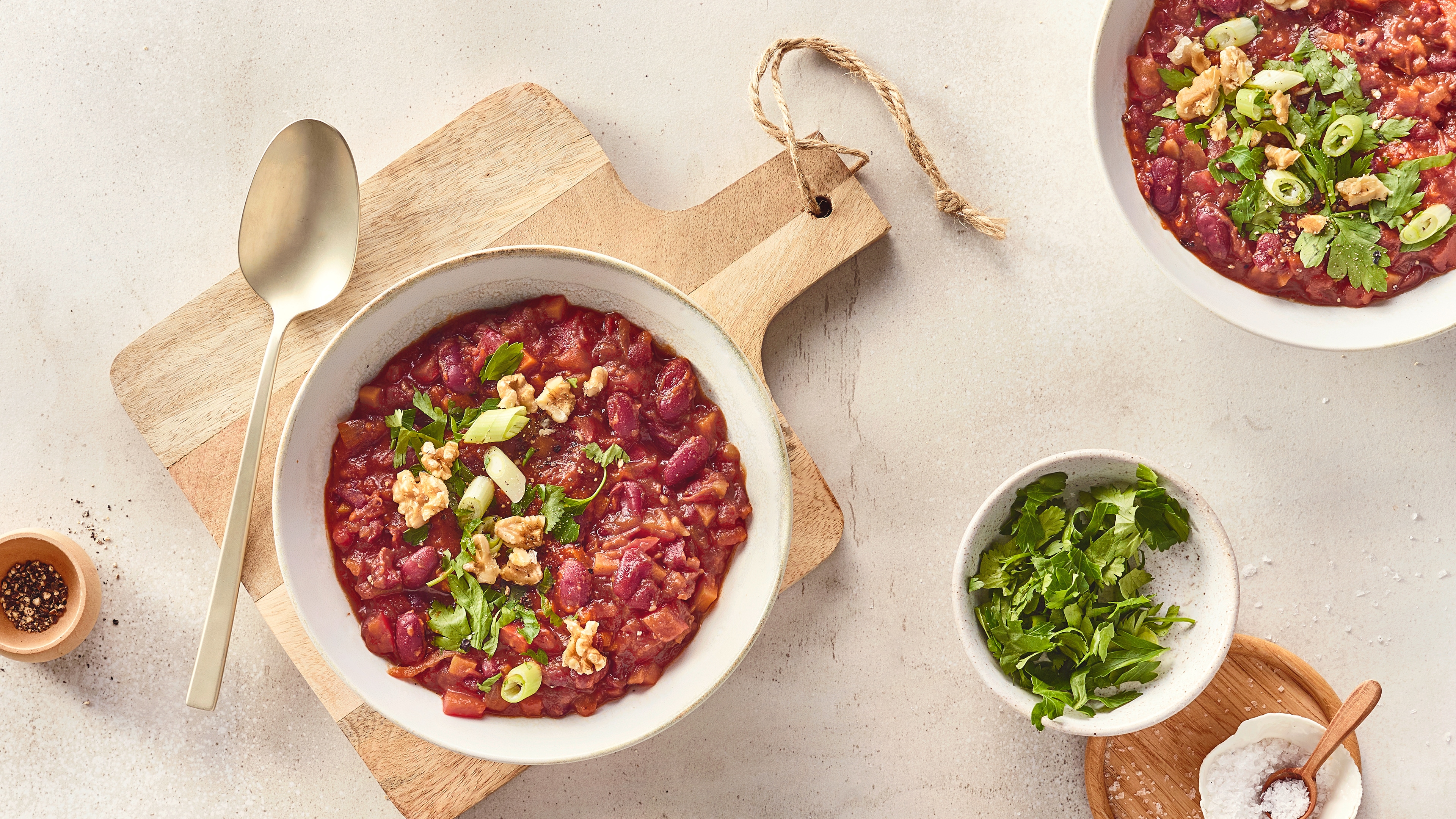 Eine Schüssel mit roter Soße, darin rote Bohnen und kleine Stücke, darauf grüne Kräuter, helle Walnüsse und Lauchstücke