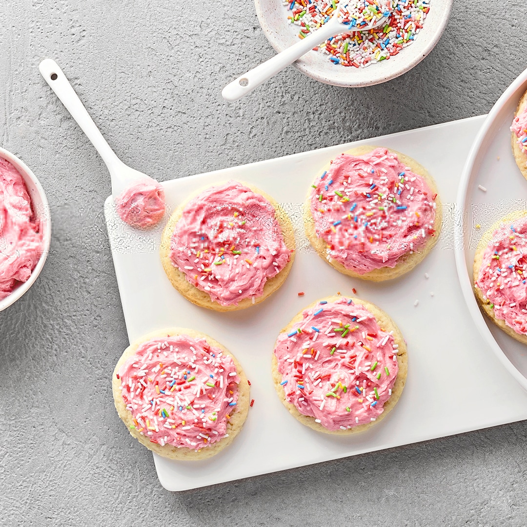 Platte mit runden Keksen, die mit einer rosa Creme und bunten Streuseln verziert sind. Daneben Schälchen mit Streuseln.