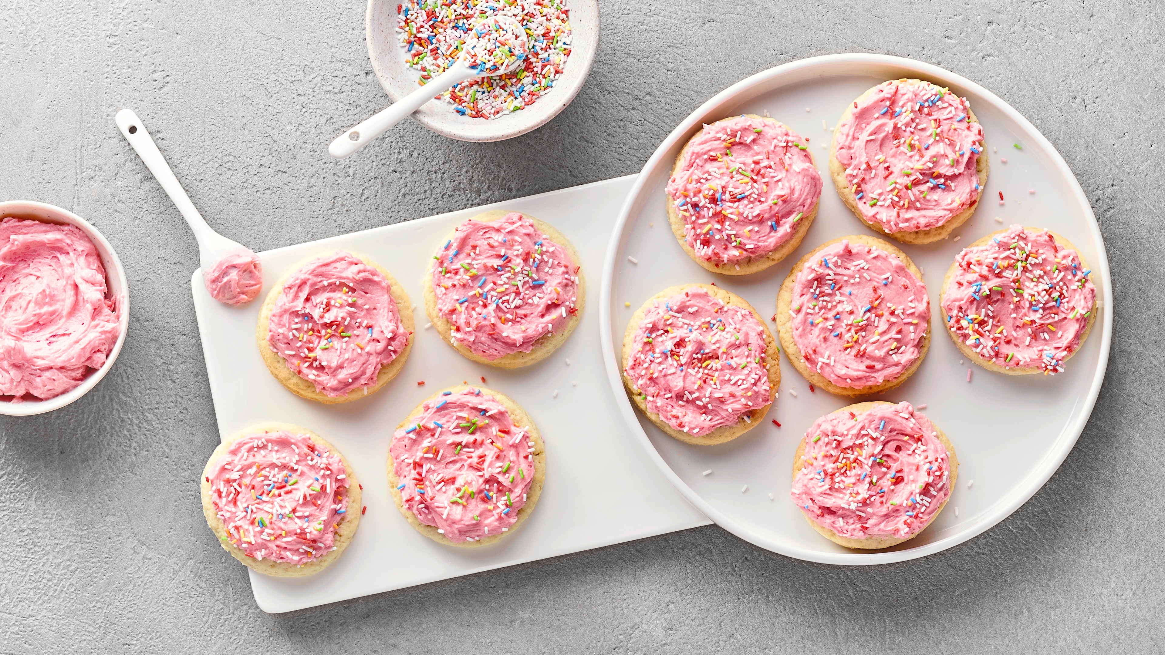 Platte mit runden Keksen, die mit einer rosa Creme und bunten Streuseln verziert sind. Daneben Schälchen mit Streuseln.
