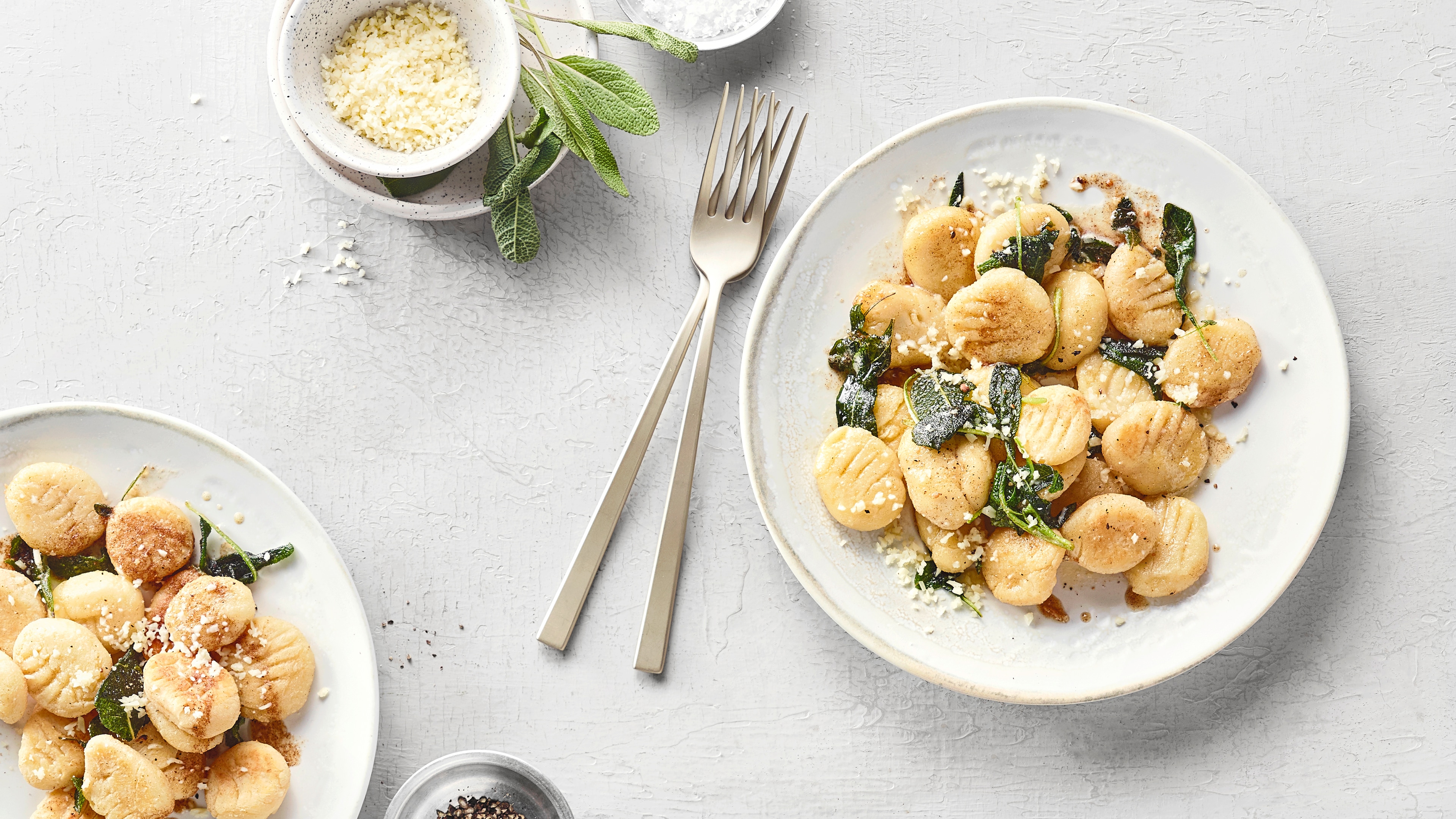 Flache Gnocchi mit Käse und grünen Salbeiblättern auf einem Teller. Daneben Besteck, ein Schälchen mit Käse und angeschnitten ein weiterer Teller.