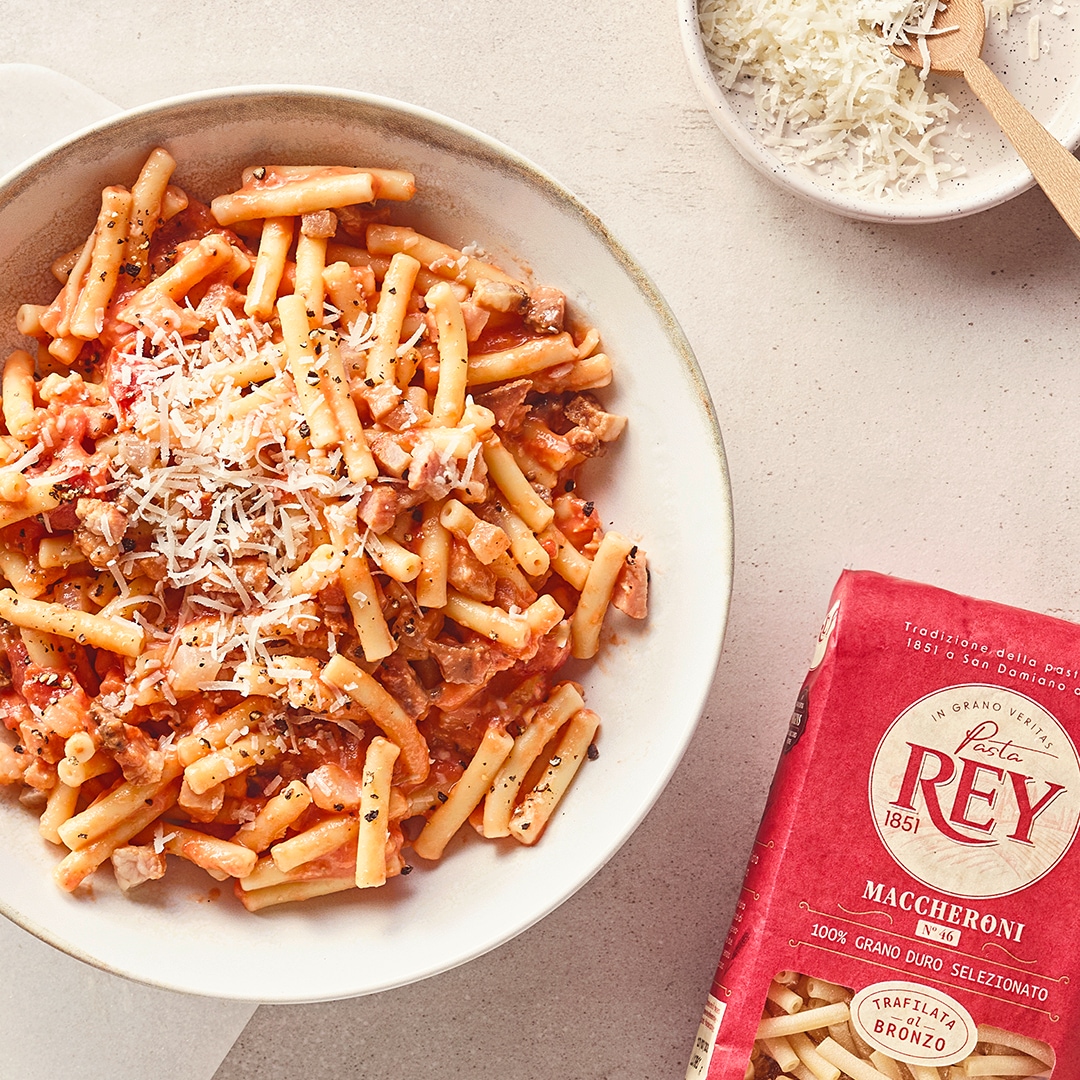 Röhrennudeln in Tomatensauce mit geriebenem Käse auf einem weißen Teller; daneben Pasta Rey Maccheroni Verpackung.