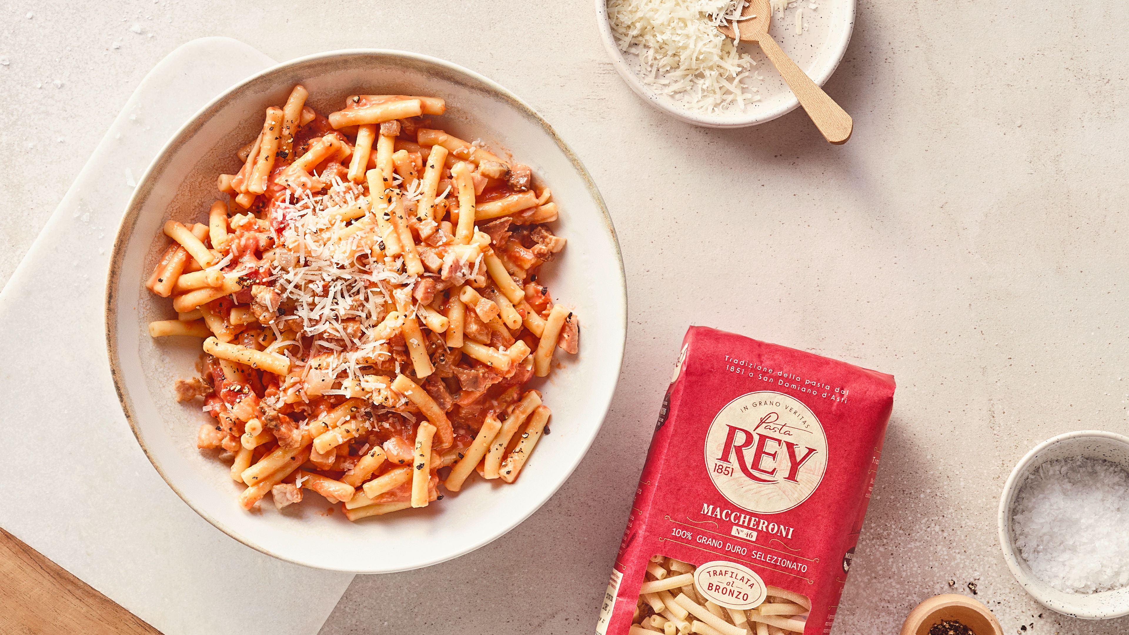 Röhrennudeln in Tomatensauce mit geriebenem Käse auf einem weißen Teller; daneben Pasta Rey Maccheroni Verpackung.