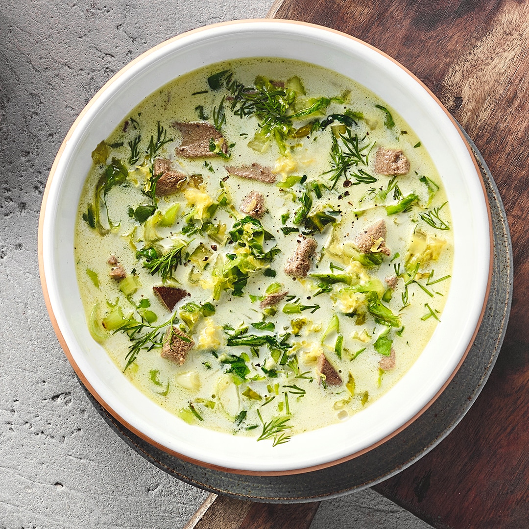 Ein Teller mit grüner cremiger Suppe, darin Fleischstücke, Kohlstreifen und Dill.