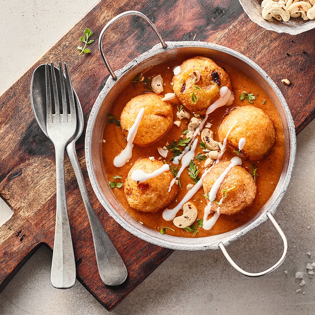 Ein gusseiserner Topf mit krossen Teigkugeln in einer hellbraunen, klaren Sauce, garniert mit Sahne und gerösteten Cashews