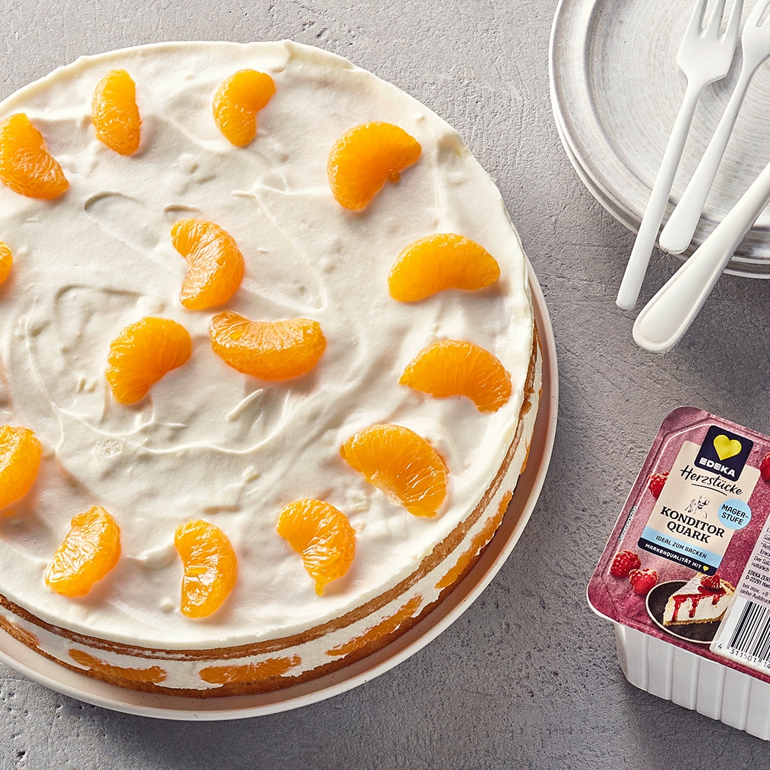 Eine Torte mit weißer Quarkschicht und Mandarinen, daneben EDEKA Herzstücke Konditor Quark