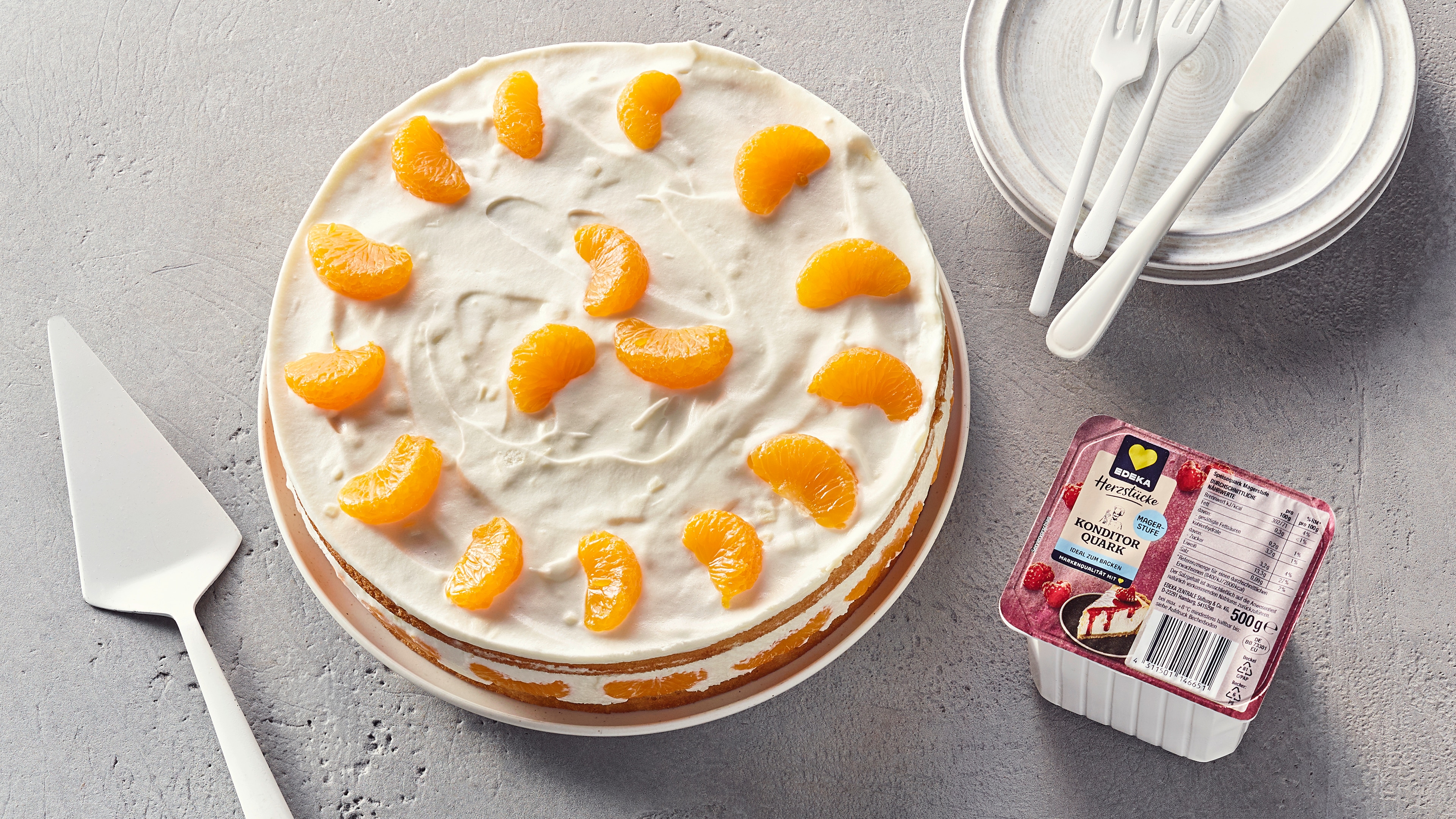 Eine Torte mit weißer Quarkschicht und Mandarinen, daneben EDEKA Herzstücke Konditor Quark