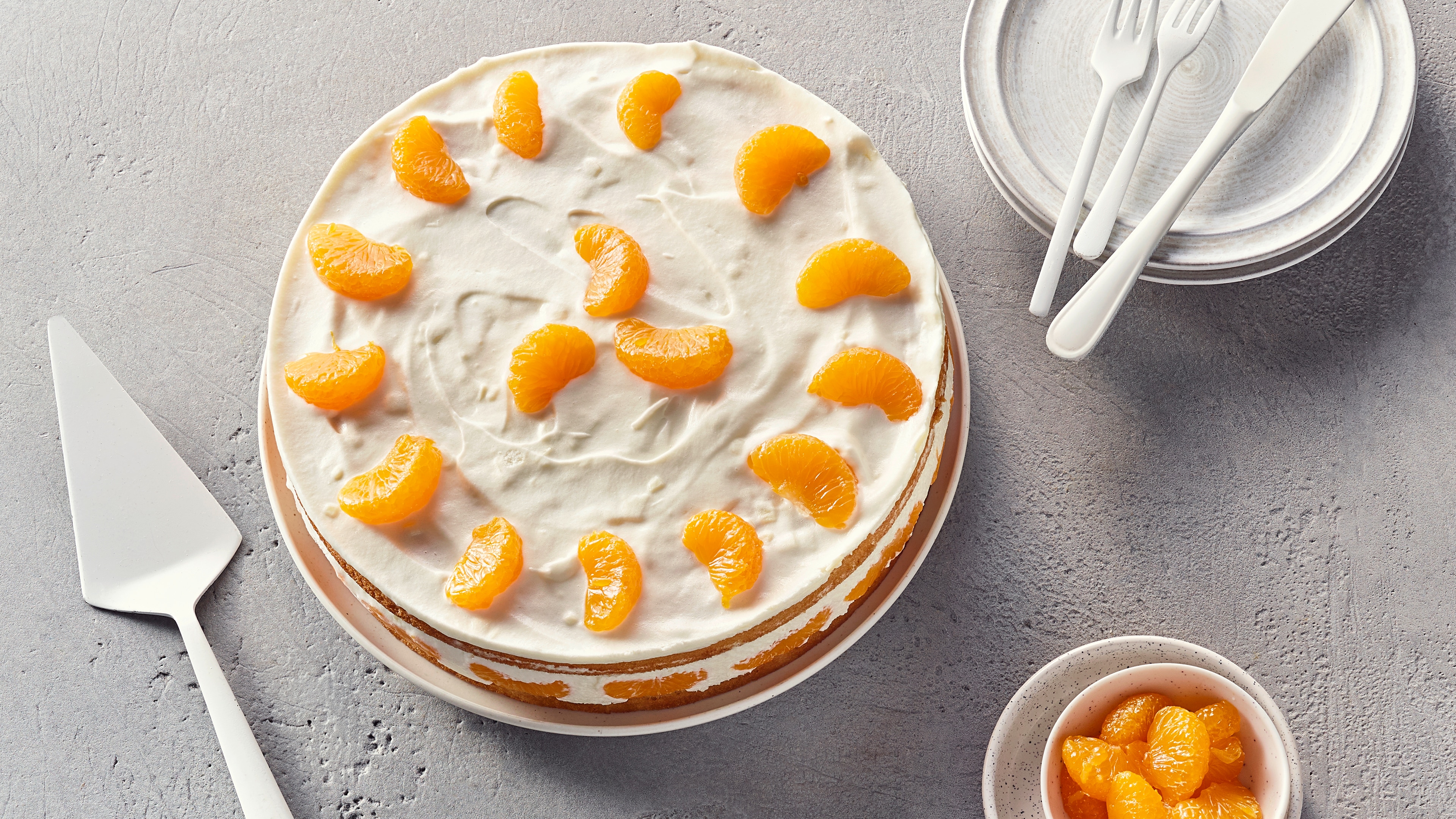 Eine Torte mit weißer Quarkschicht und Mandarinen, daneben kleine Teller und ein Schälchen mit Manarinen