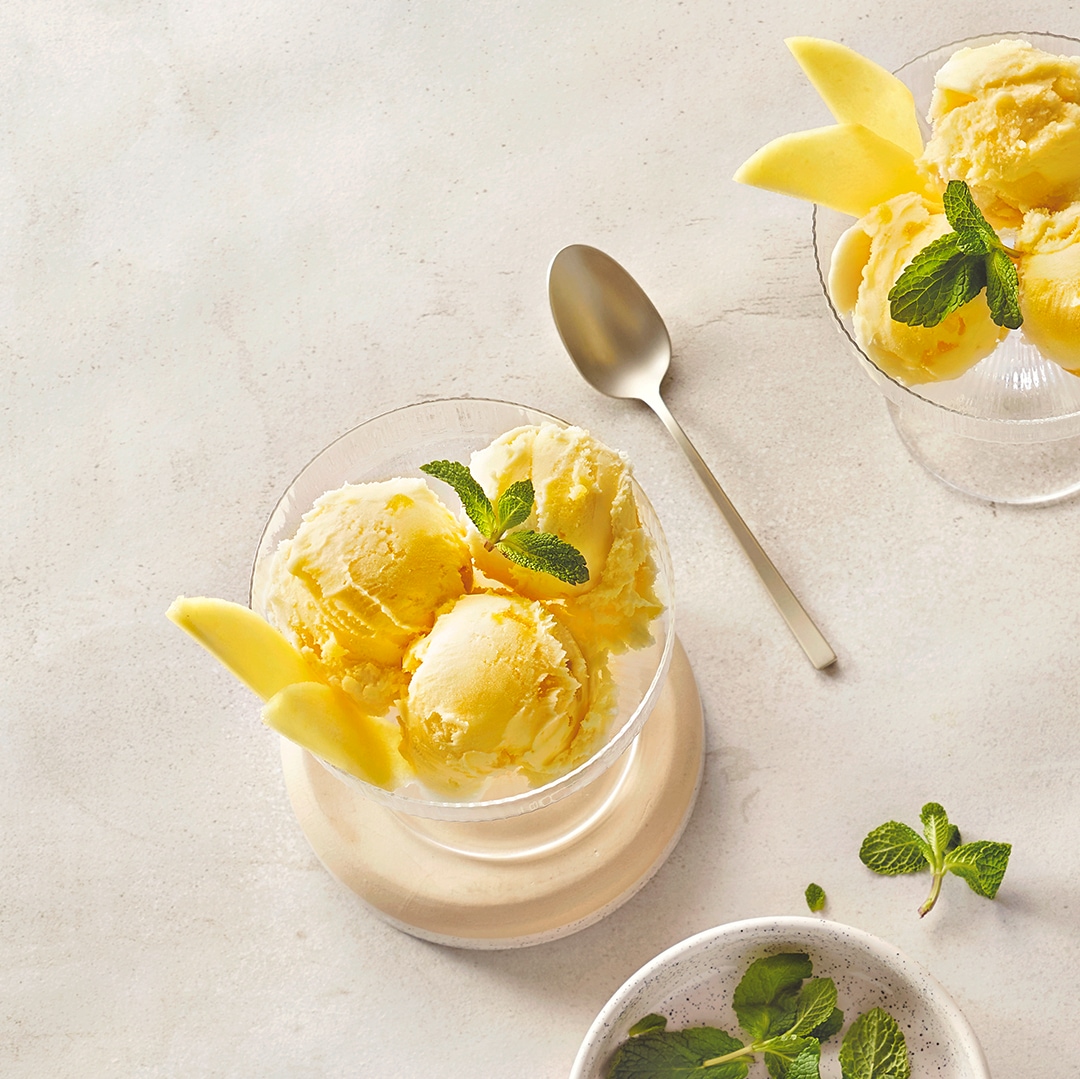 Leuchtend gelbe Eiskugeln in Glas mit Mangospalten daneben