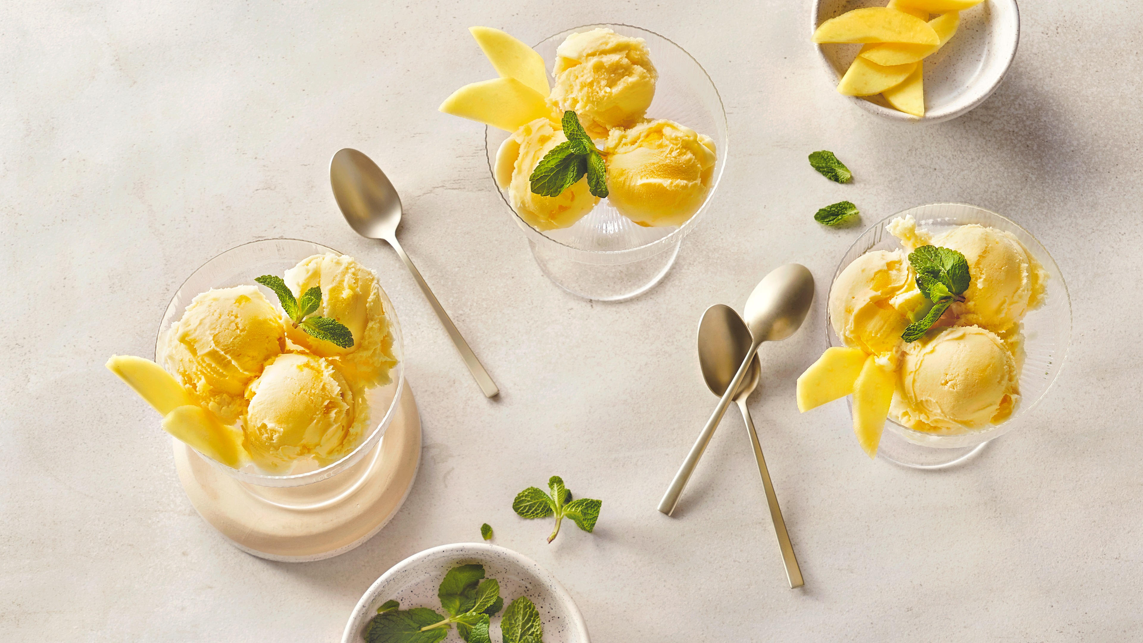 Leuchtend gelbe Eiskugeln in Gläsern mit Mangospalten daneben