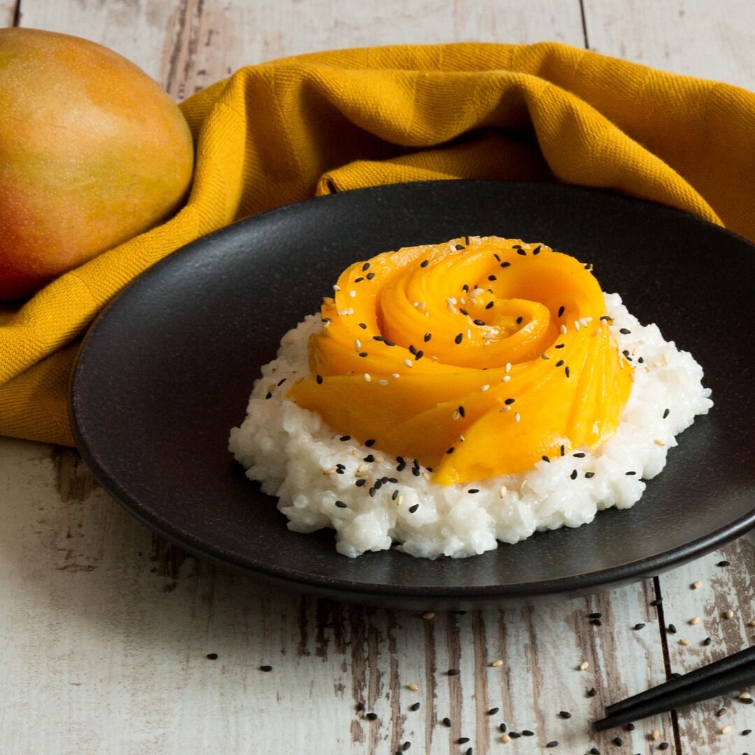 Ein Teller mit Reis, getoppt mit Mangoscheiben und schwarzen Sesam, daneben eine Mango