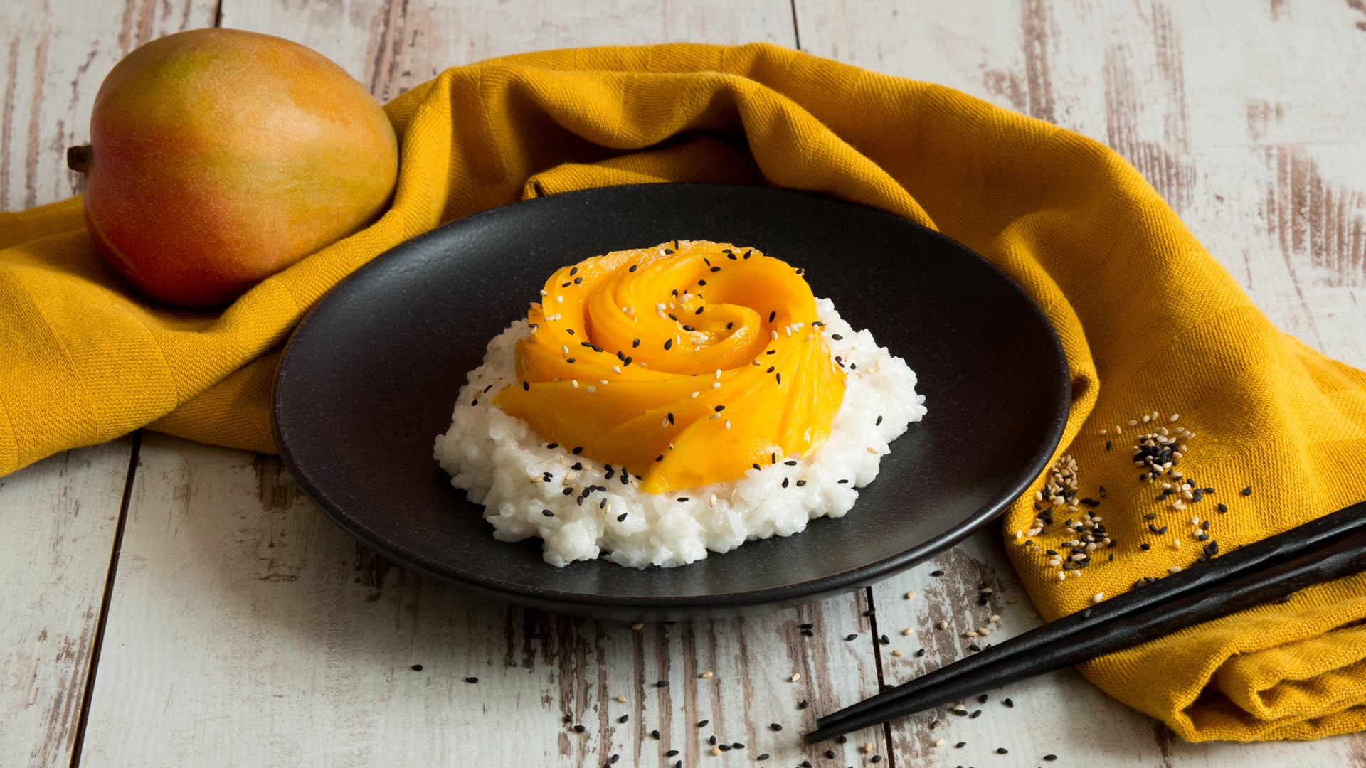 Ein Teller mit Reis, getoppt mit Mangoscheiben und schwarzen Sesam, daneben eine Mango