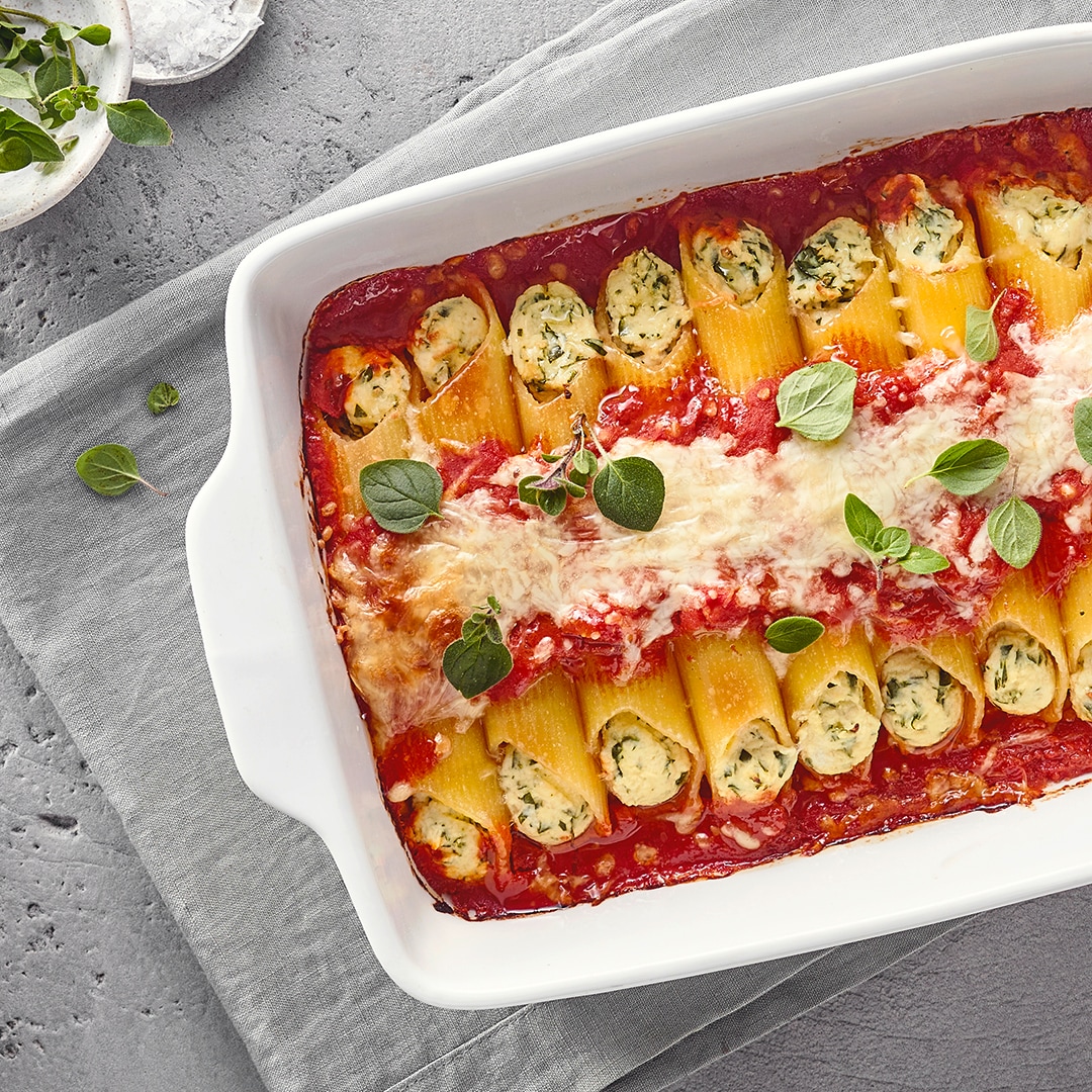 Eine Form mit gebackenen Nudelrollen, gefüllt mit Ricotta und Kräutern, bedeckt mit Tomatensoße, Käse und frischem Basilikum