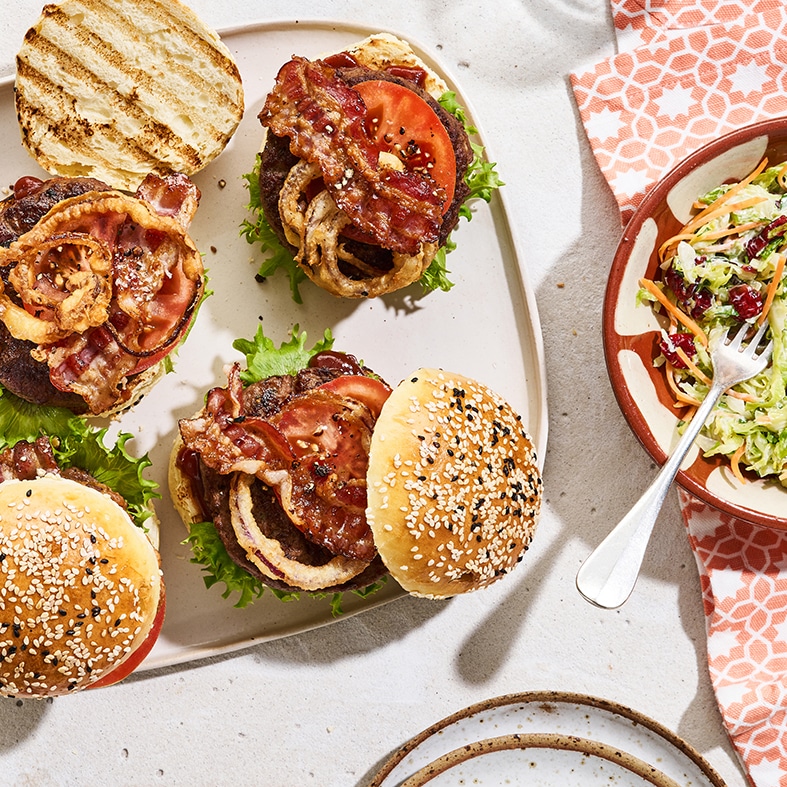 Teller mit drei aufgeklappten Burgern, belegt mit glasiertem Bacon, Tomaten, Zwiebeln und Salat. Rechts daneben steht eine Schale mit buntem Weißkohlsalat, in der eine Gabel liegt.