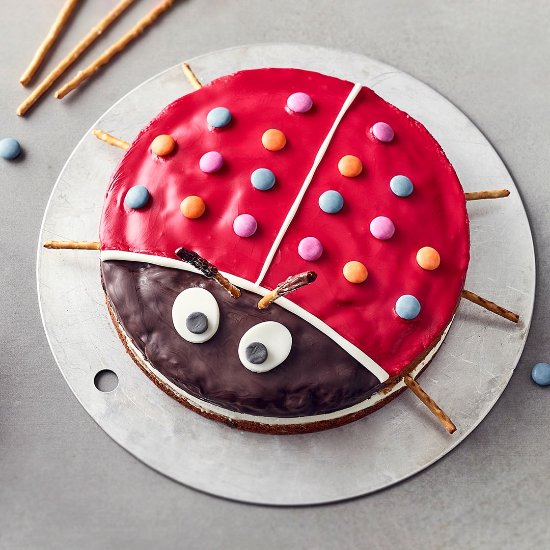 Ein Kuchen, dekoriert wie ein Marienkäfer mit roter Glasur, Smarties als Punkten, Fondant-Augen und Salzstangenbeinen