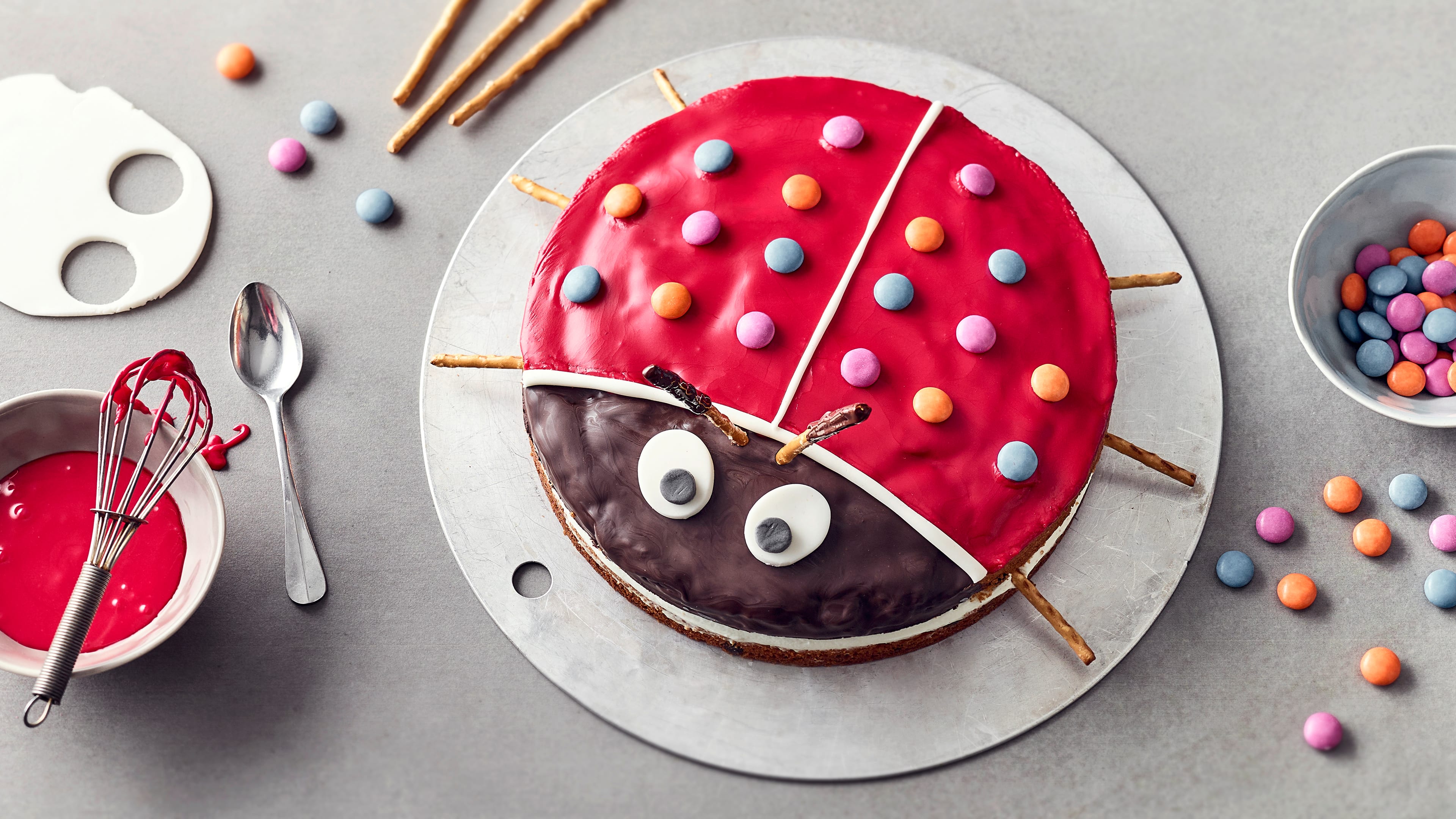 Rezept für Marienkäfer-Kuchen Ein Kuchen, dekoriert wie ein Marienkäfer mit roter Glasur, Smarties als Punkten, Fondant-Augen und Salzstangenbeinen