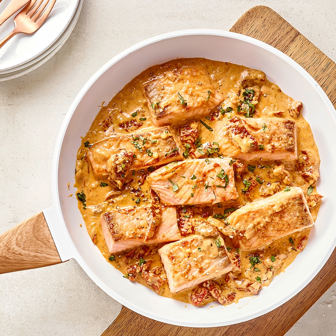 Gebratene Lachsstücke in cremiger Sauce in einer Pfanne, serviert auf einem Holzbrett mit Gewürzen.