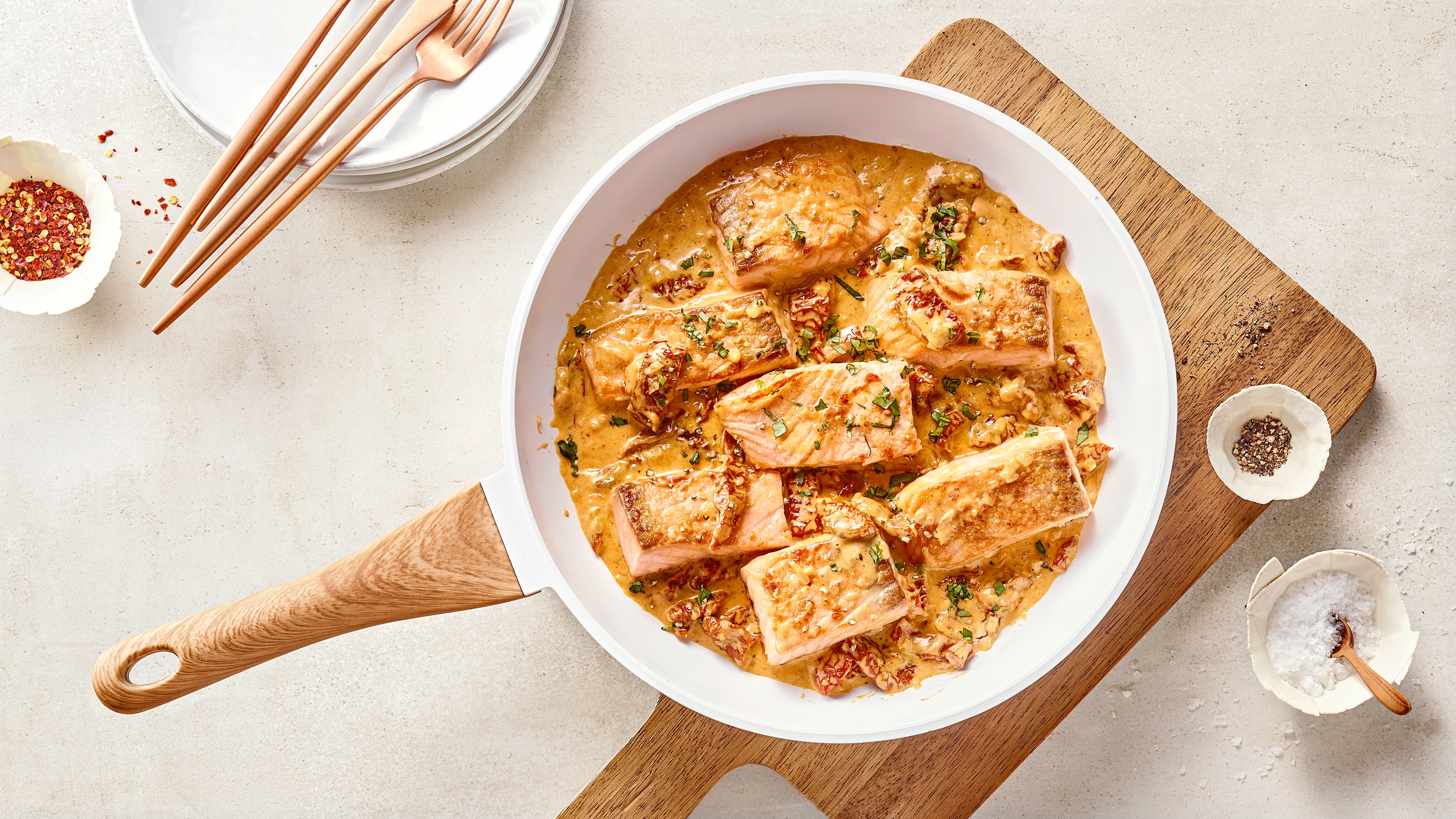 Gebratene Lachsstücke in cremiger Sauce in einer Pfanne, serviert auf einem Holzbrett mit Gewürzen.