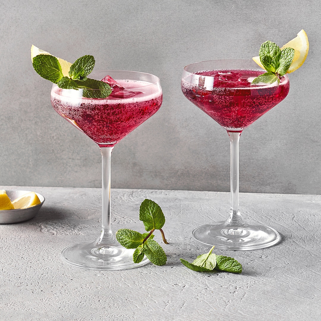 Zwei Gläser mit violettem Getränk, darin Eiswürfel, jeweils ein Zitronenschnitz und grüne Minzblätter am Rand.