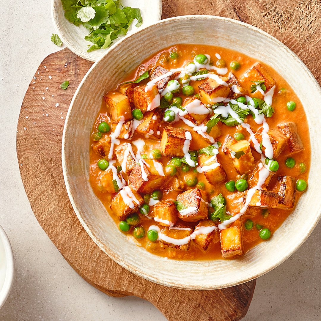 Eine Schale mit orangefarbener Currysoße, darin goldbraun gebratene Paneerwürfel und grüne Erbsen, garniert mit weißen Cremestreifen