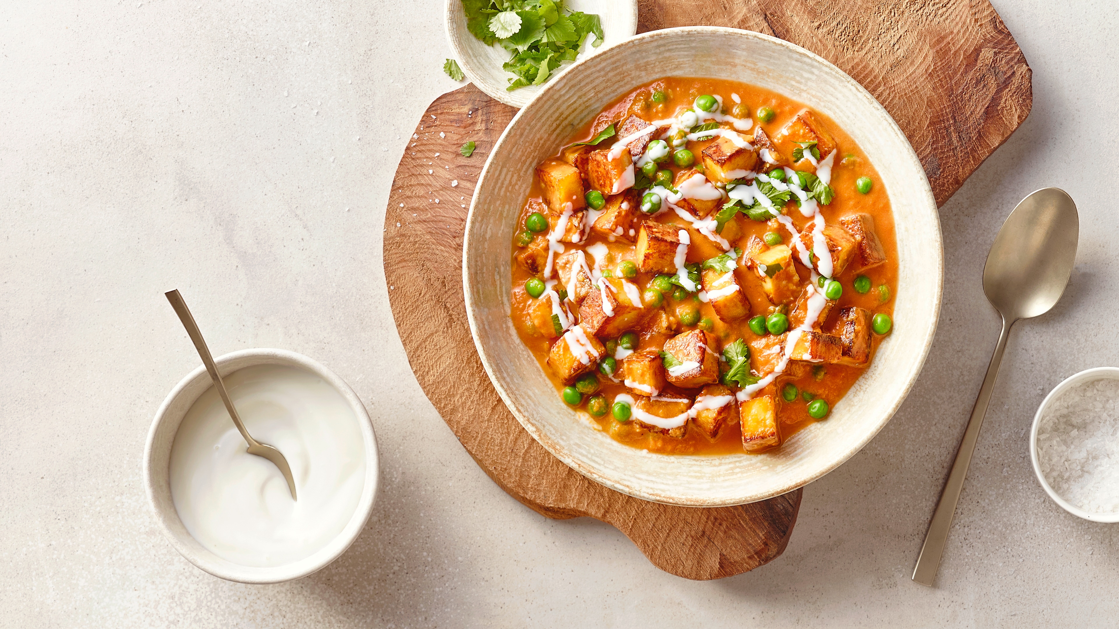 Eine Schale mit orangefarbener Currysoße, darin goldbraun gebratene Paneerwürfel und grüne Erbsen, garniert mit weißen Cremestreifen