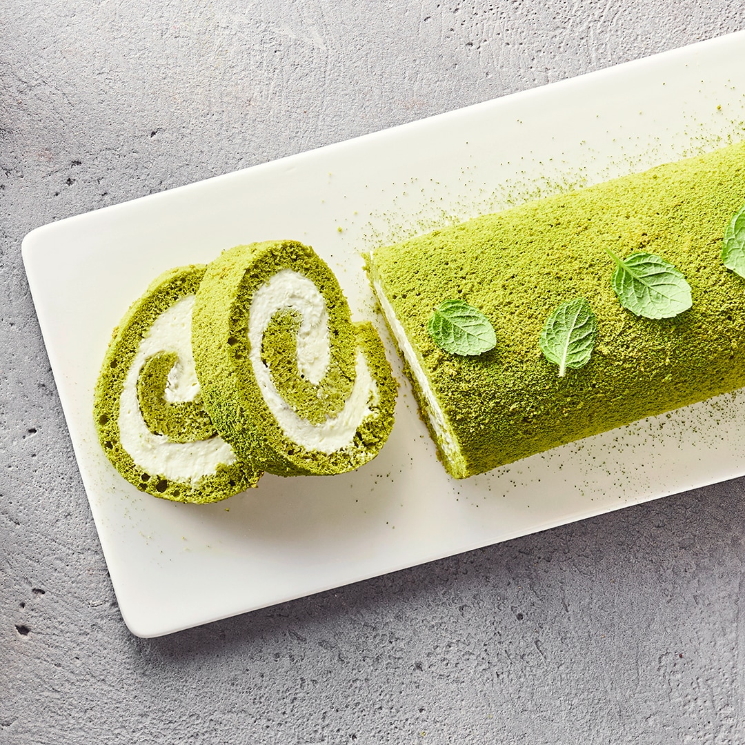 Ein Teller mit grünem Matcha-Rollenkuchen, bestreut mit Matchapulver, gefüllt mit weißer Creme, daneben zwei angeschnittene Stücke