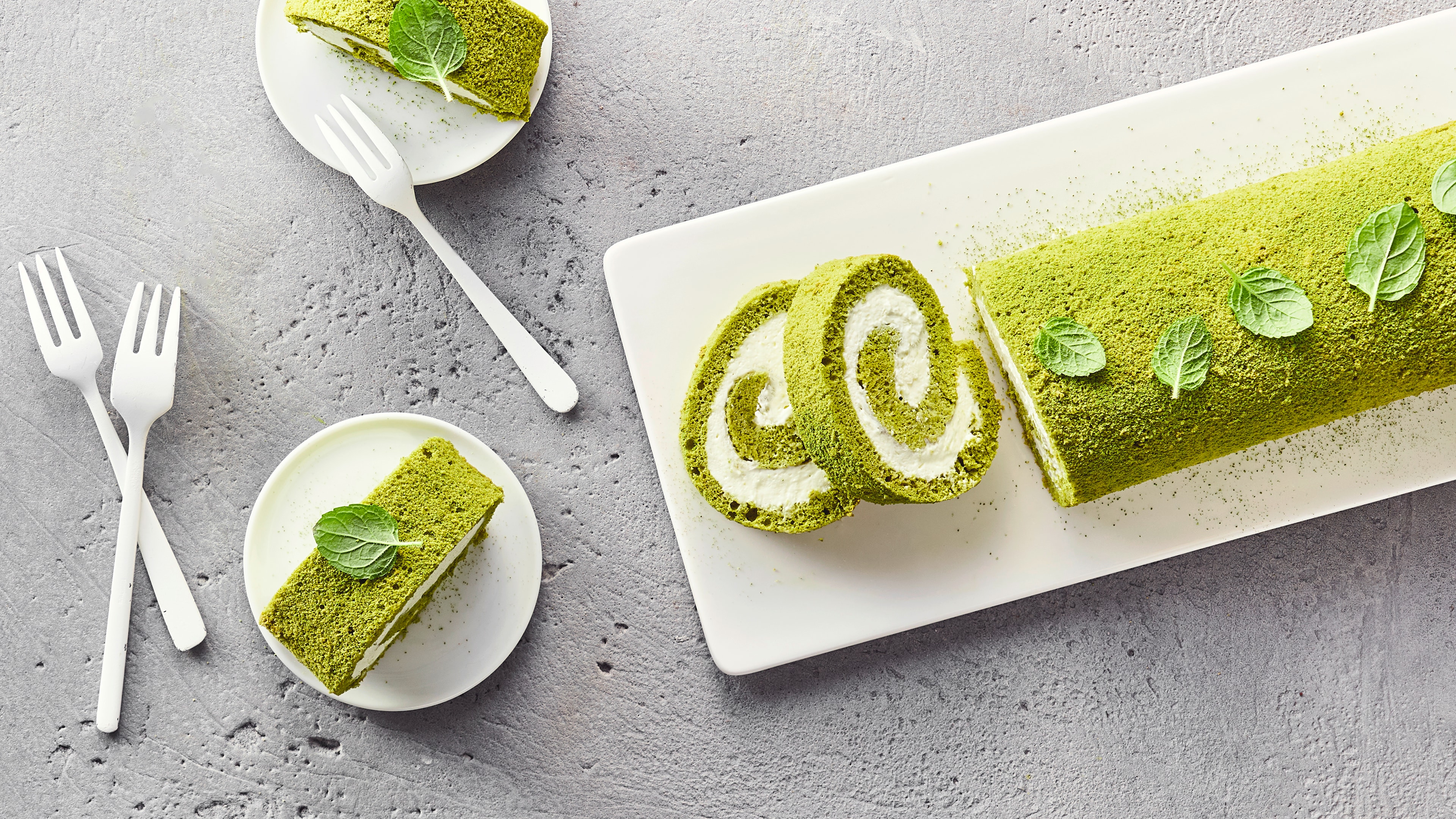 Rezept für Matcha Biskuitrolle Ein Teller mit grünem Matcha-Rollenkuchen, bestreut mit Matchapulver, gefüllt mit weißer Creme, daneben zwei angeschnittene Stücke