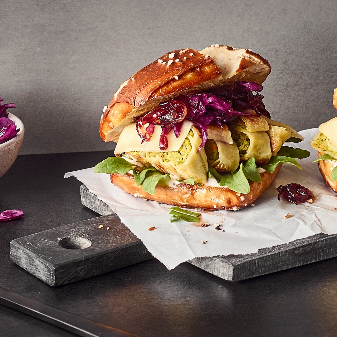 Laugen-Burger, gefüllt mit Rotkohl, Käse und Gemüse-Maultaschen, Salat und heller Sauce, auf einem Steinbrett