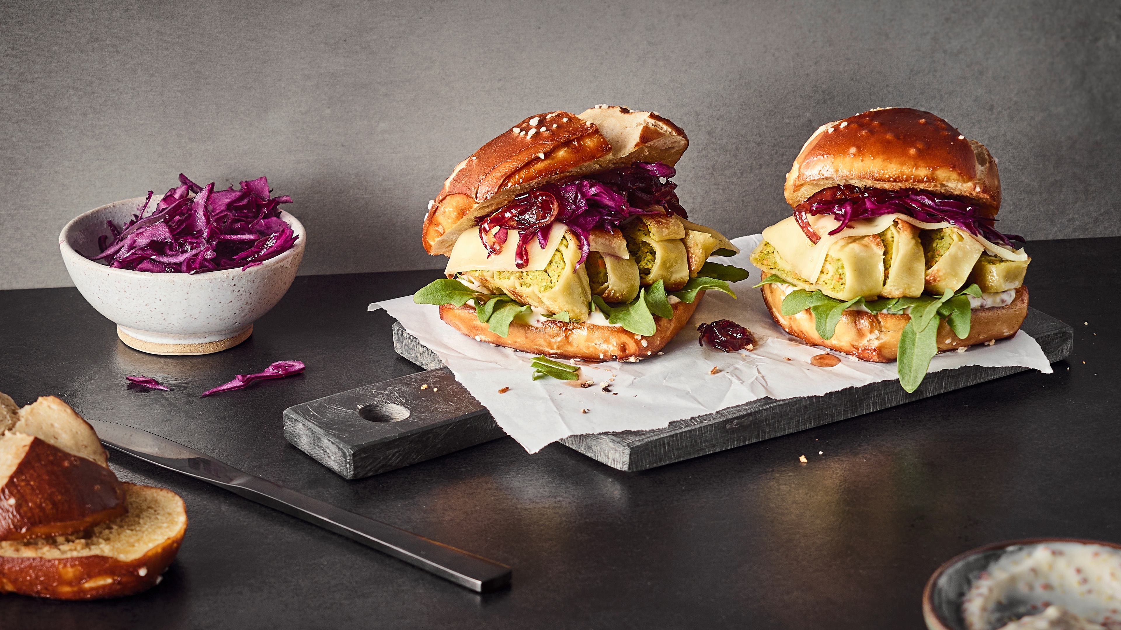 Rezept für Maultaschen-Burger Laugen-Burger, gefüllt mit Rotkohl, Käse und Gemüse-Maultaschen, Salat und heller Sauce, auf einem Steinbrett