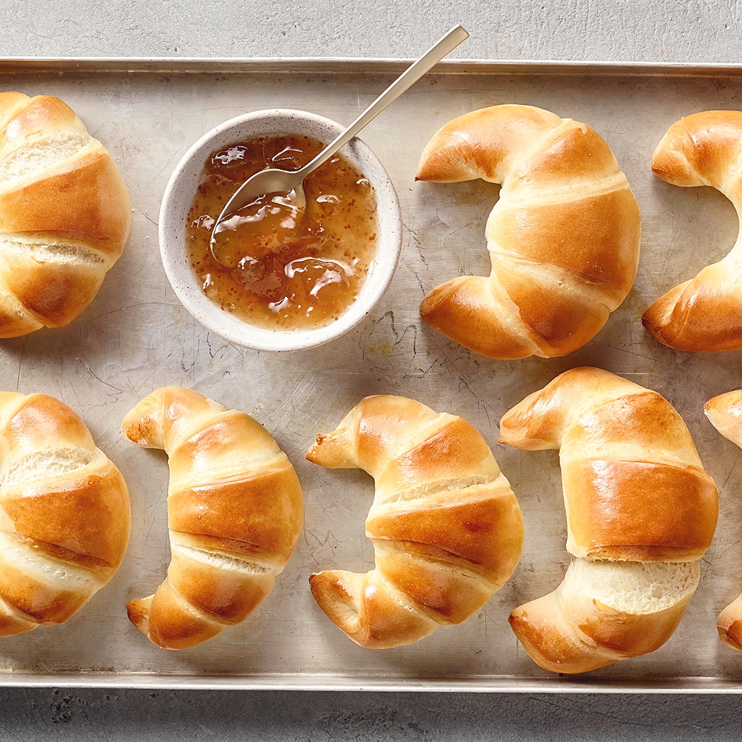 Ein Blech mit acht goldbraun gebackenen Croissants und einer Schale heller Marmelade mit Löffel.