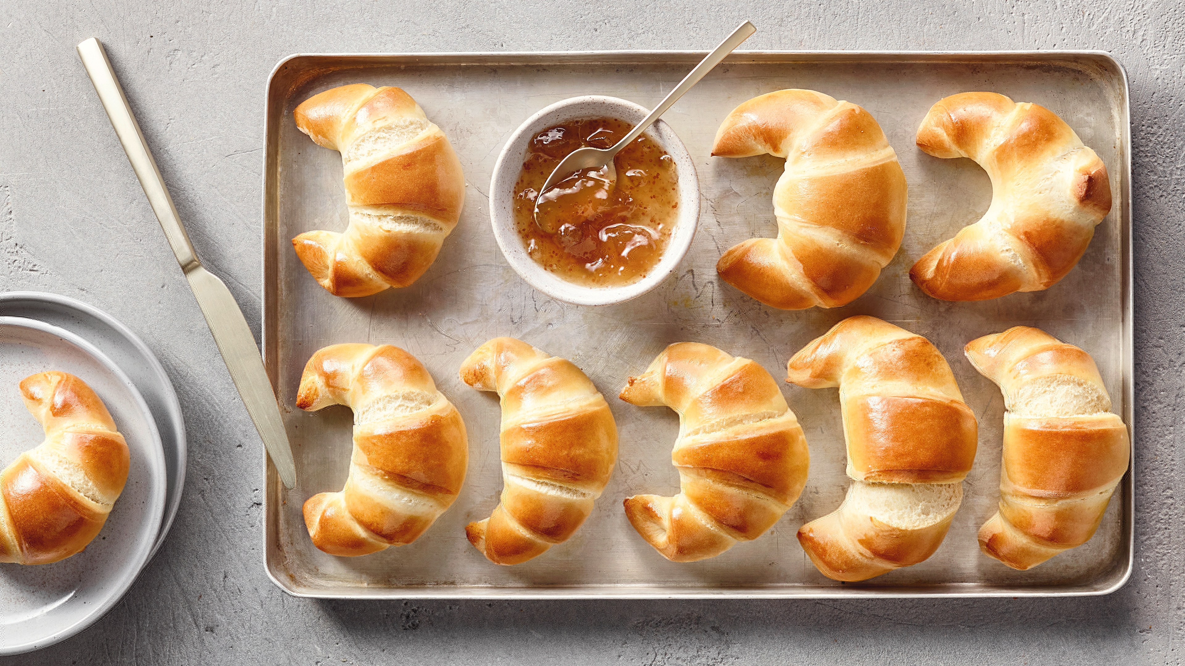 Ein Blech mit acht goldbraun gebackenen Croissants und einer Schale heller Marmelade mit Löffel.
