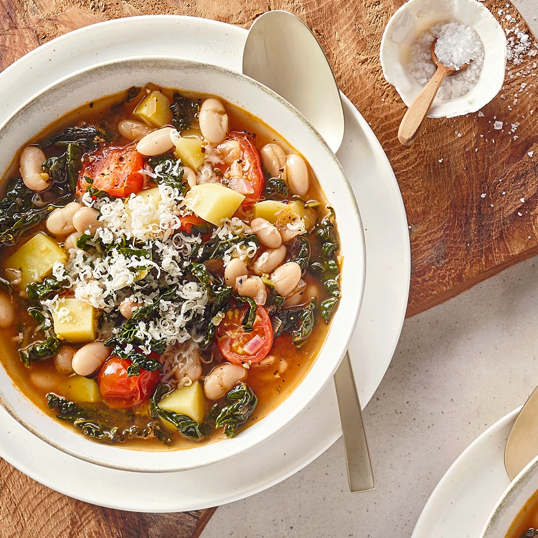 Eine Schüssel mit klarer Suppe, darin weiße Bohnen, rote Tomatenstücke, gelbe Kartoffelwürfel, dunkle Blattstücke und geriebener Käse.