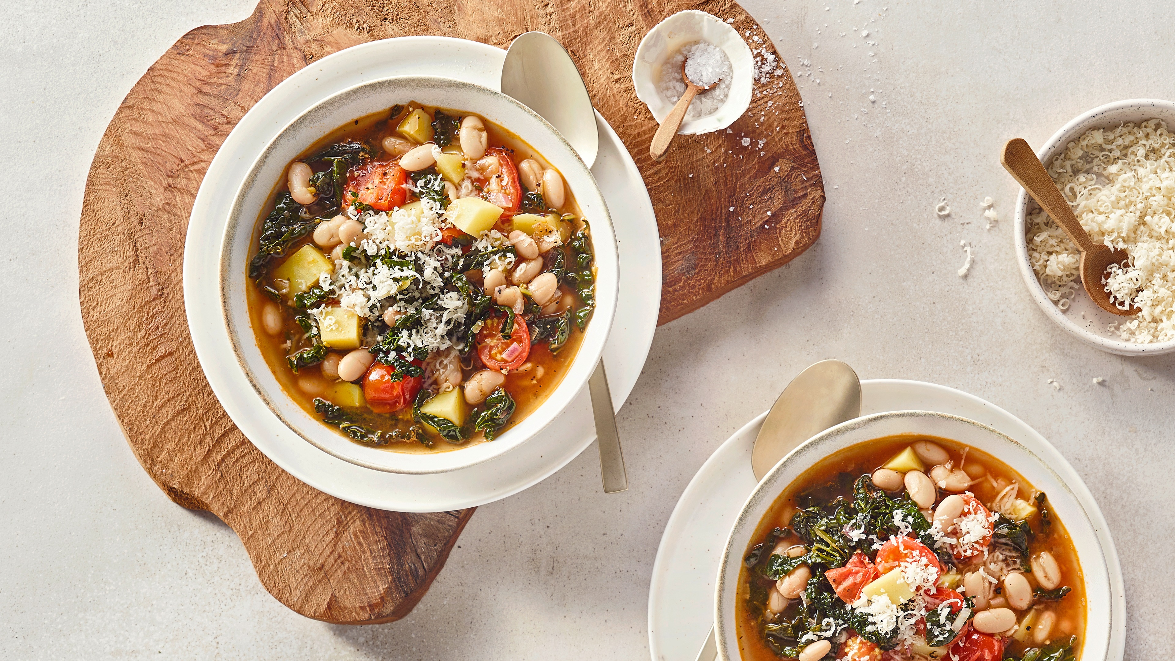 Eine Schüssel mit klarer Suppe, darin weiße Bohnen, rote Tomatenstücke, gelbe Kartoffelwürfel, dunkle Blattstücke und geriebener Käse.