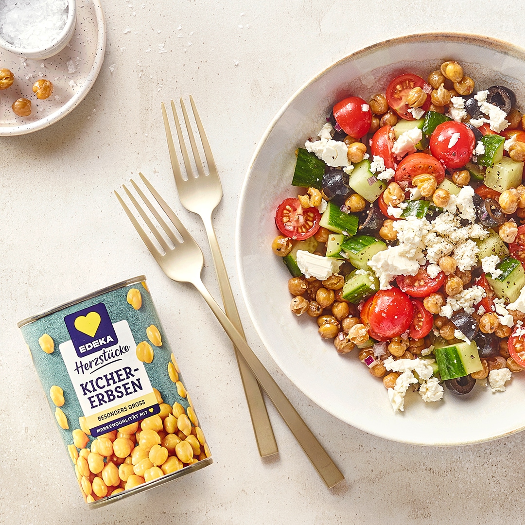Bunte Kichererbsen-Salat-Bowl mit Tomaten, Gurken, Oliven und Feta, daneben eine Dose EDEKA Herzstücke Kichererbsen und Besteck.