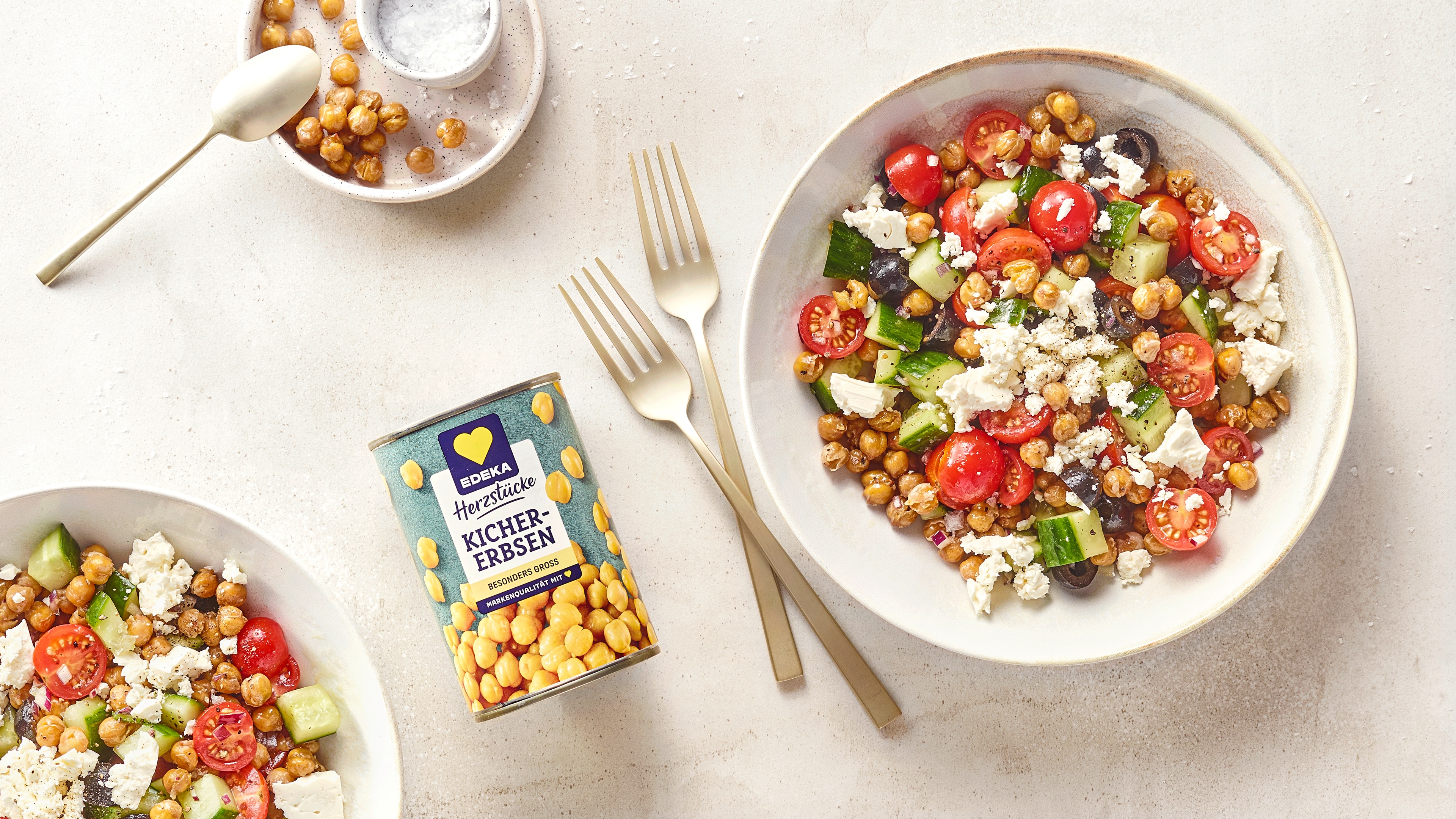 Bunte Kichererbsen-Salat-Bowl mit Tomaten, Gurken, Oliven und Feta, daneben eine Dose EDEKA Herzstücke Kichererbsen und Besteck.