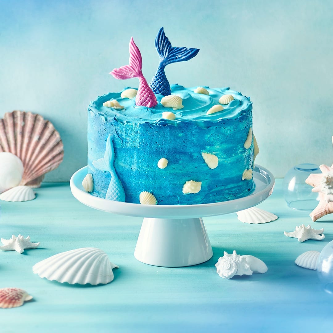 Eine blaue Creme-Torte, dekoriert mit weißen Muscheln und farbigen Meerjungfrauenflossen, auf einer Torten-Etagere