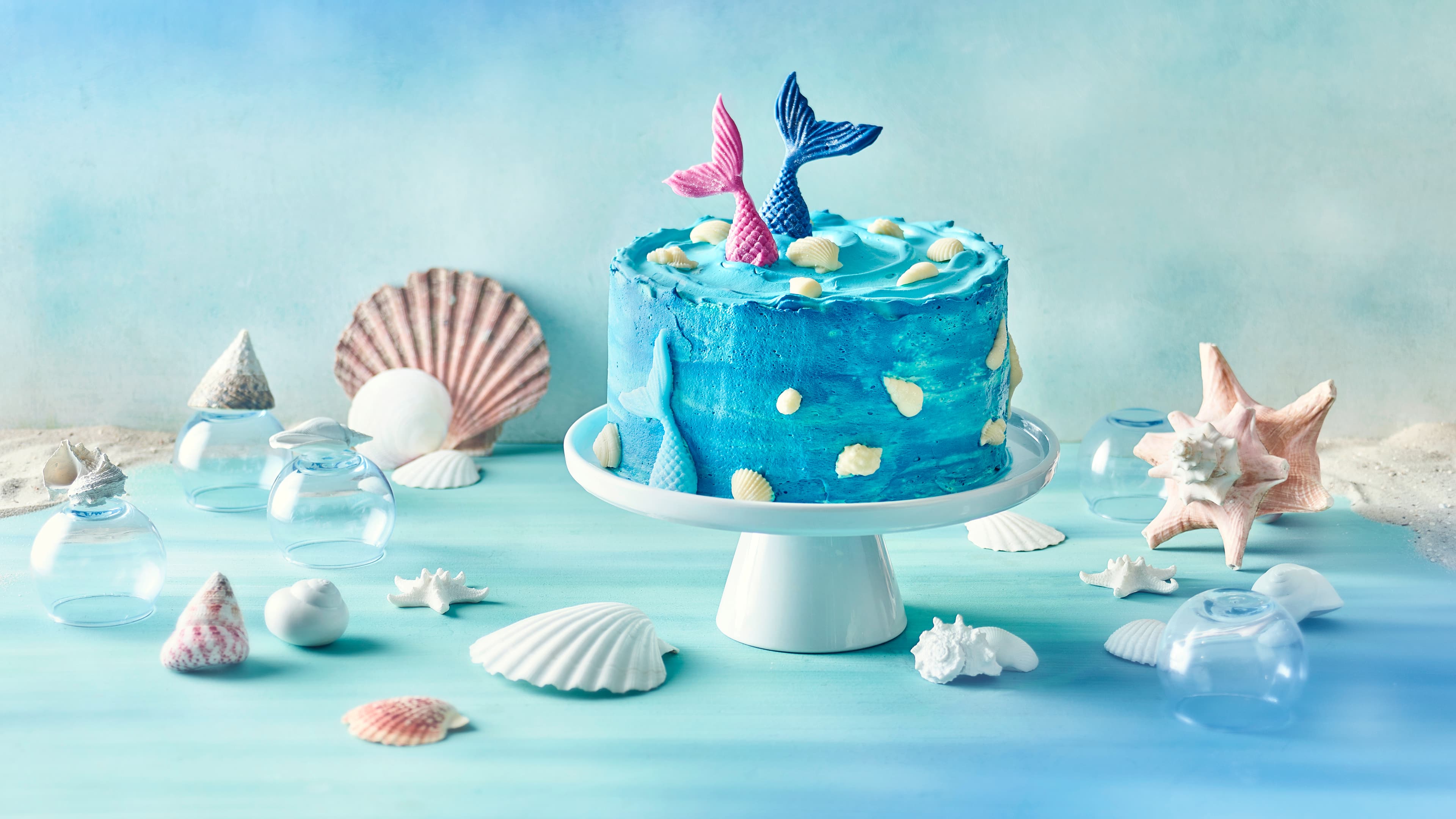 Rezept für Meerjungfrauen-Torte Eine blaue Creme-Torte, dekoriert mit weißen Muscheln und farbigen Meerjungfrauenflossen, auf einer Torten-Etagere