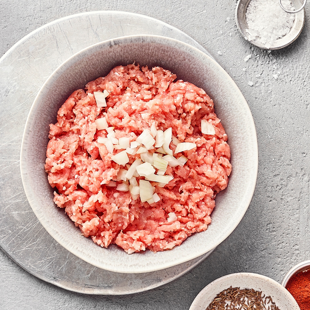Schale mit rohem, durchgedrehtem und gewürztem Schweinefleisch mit Zwiebeln. Oben rechts und unten rechts kleine Schüsseln mit Rosmarin, Paprikapulver, Salz und Zwiebeln.