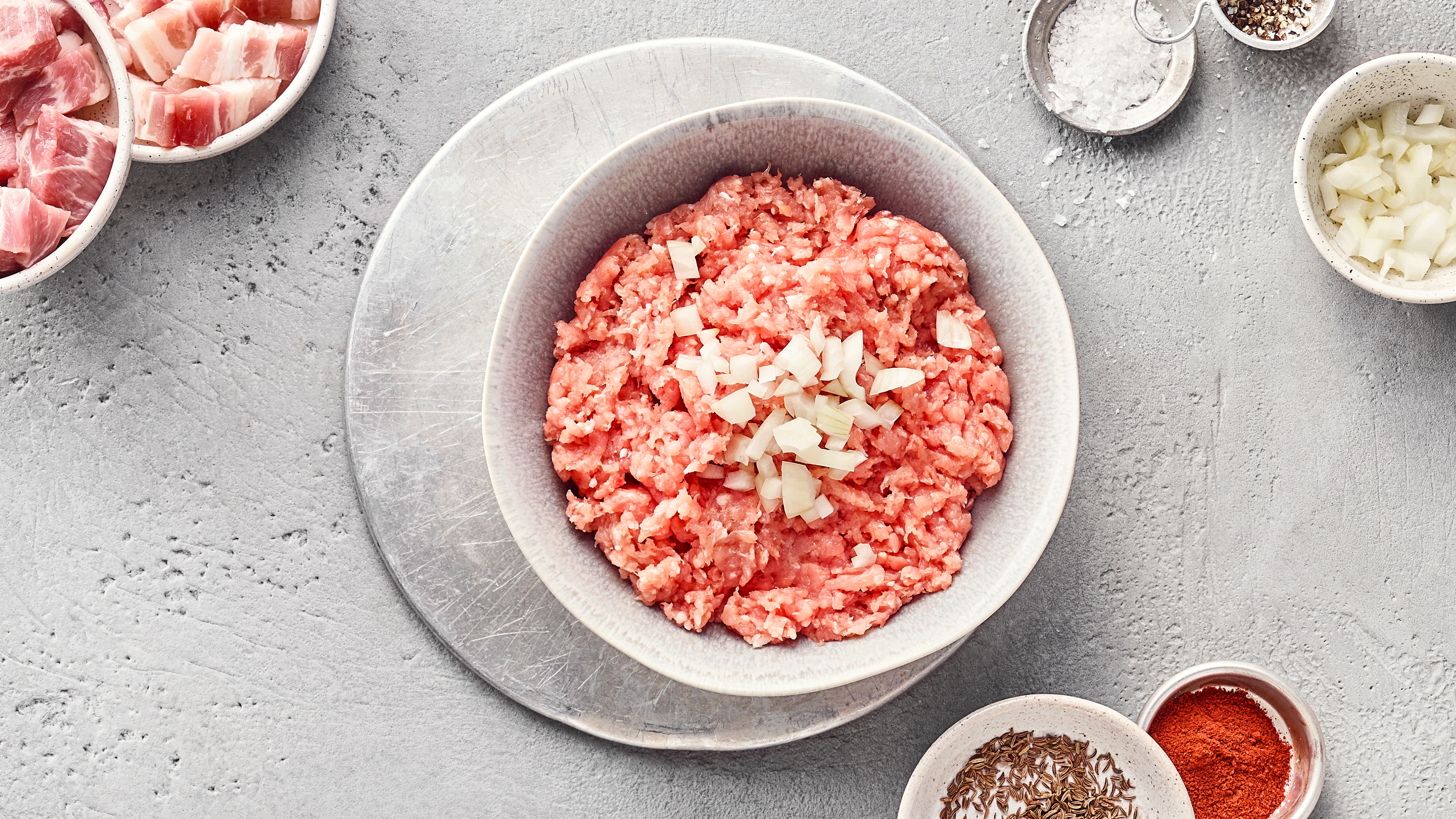 Schale mit rohem, durchgedrehtem und gewürztem Schweinefleisch mit Zwiebeln. Oben links stehen Schüsseln mit Fleischwürfeln, oben rechts und unten rechts kleine Schüsseln mit Rosmarin, Paprikapulver, Salz und Zwiebeln.