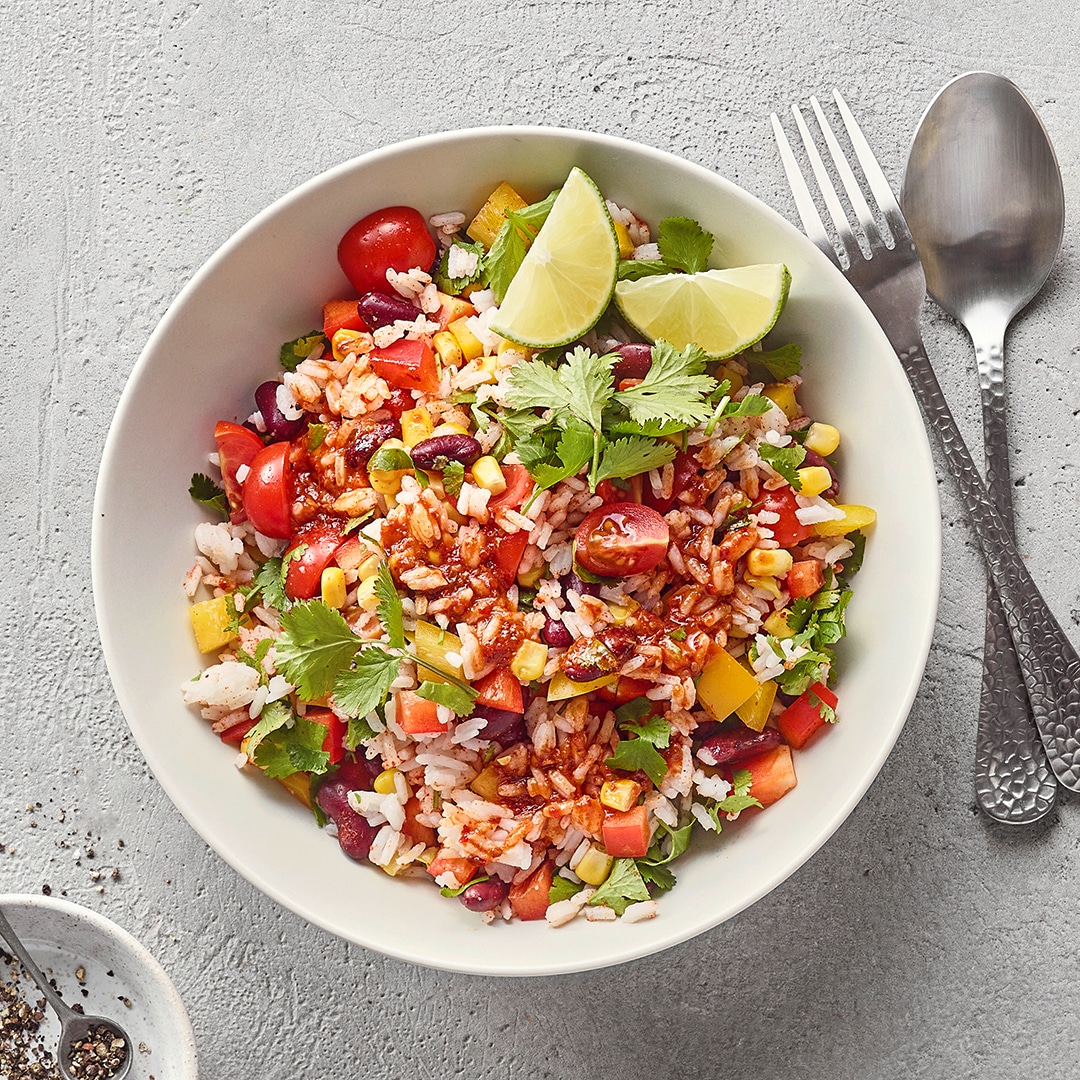 Eine Schüssel mit Reis, roten Bohnen, Mais, Paprikastücken, Tomatenhälften, frischen Kräutern und Limettenspalten.